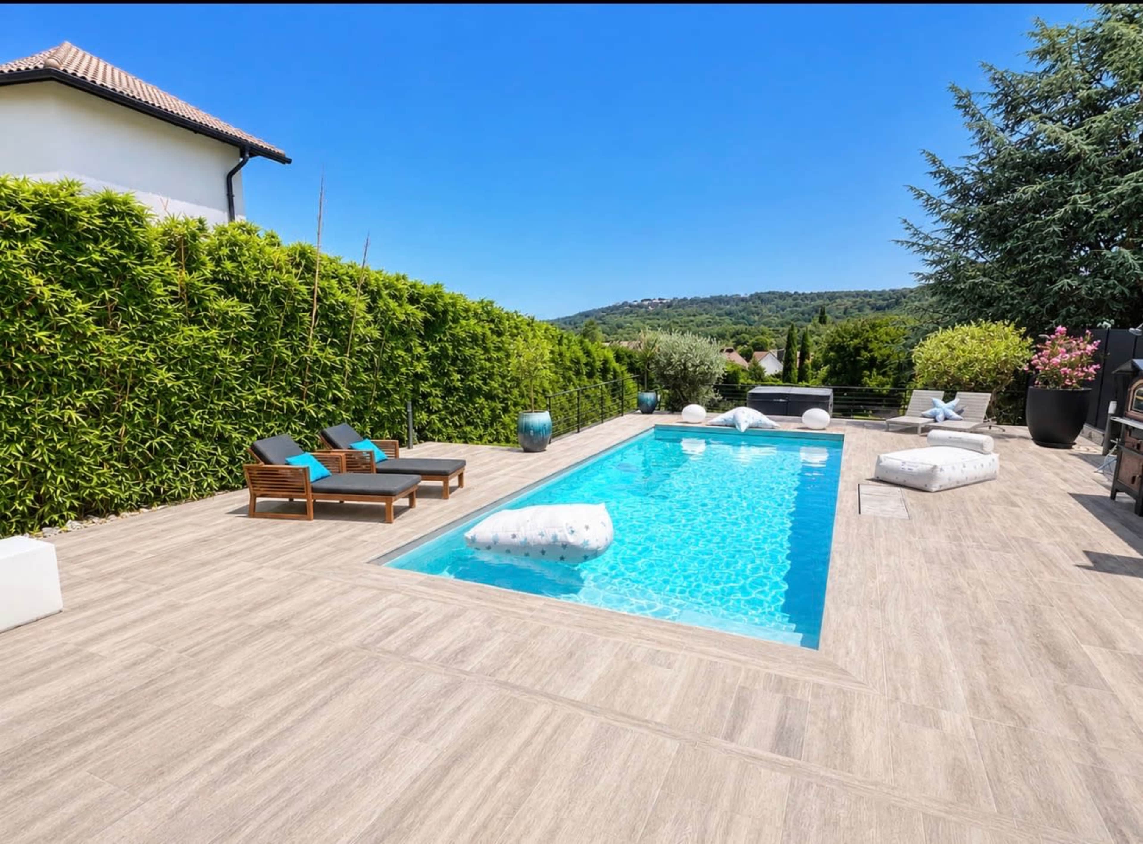 The image shows a modern backyard with a swimming pool surrounded by wooden lounge chairs and white cushions, enclosed by lush greenery.