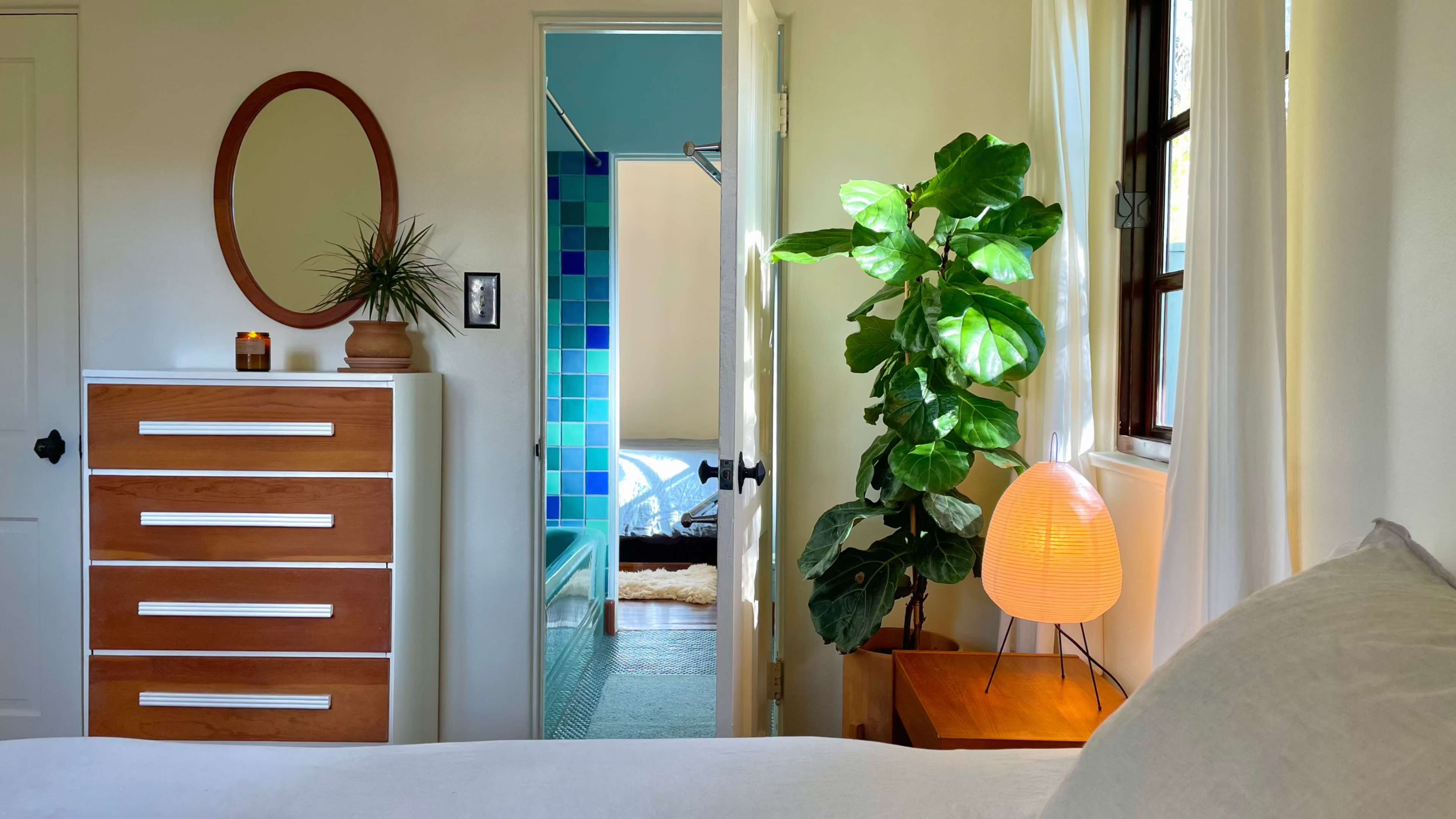 The scene shows a bright, tidy bedroom with a dresser, a potted plant, and a glowing lamp, leading to a bathroom with colorful tiles visible through an open door.