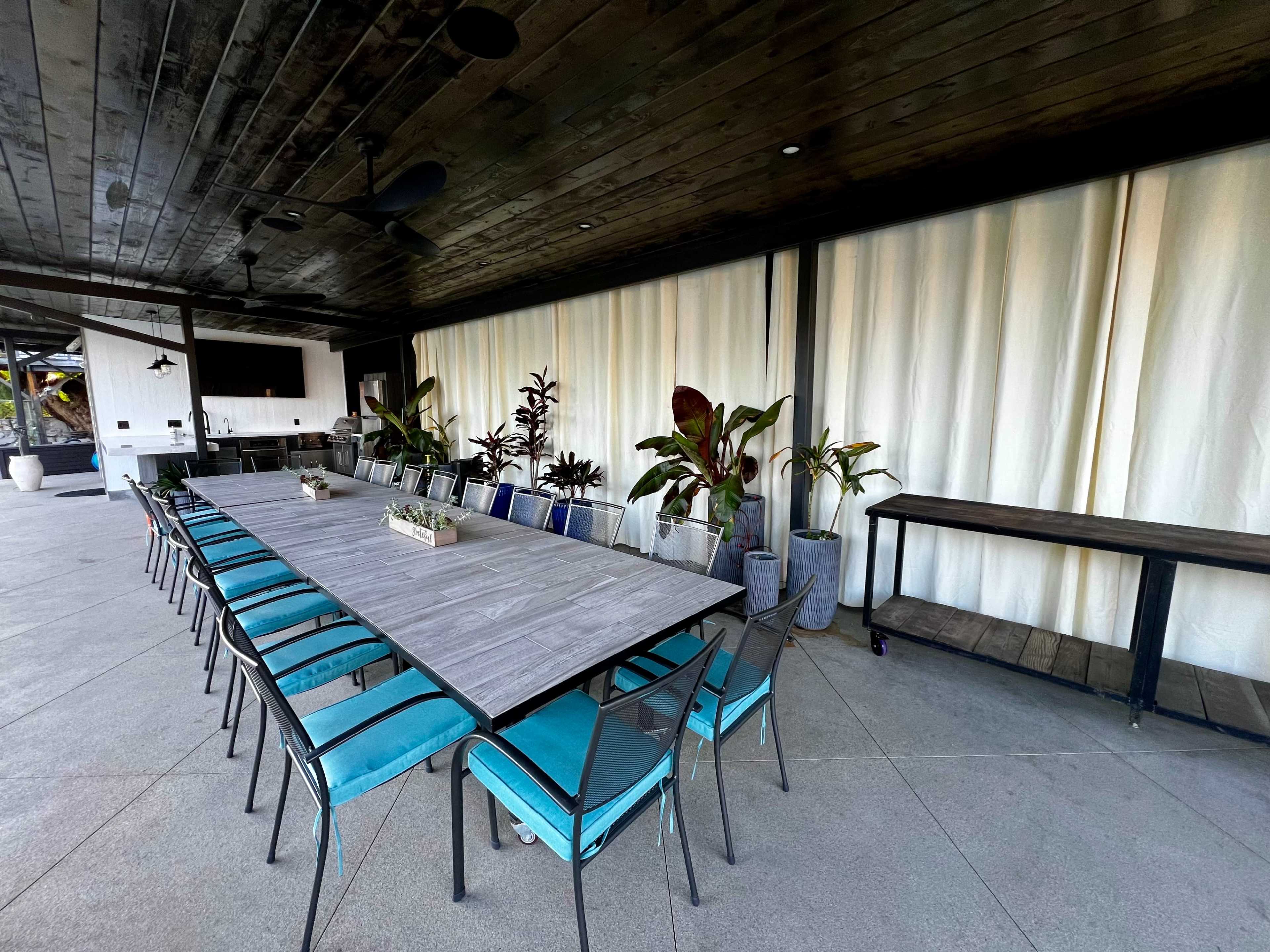 A long, rectangular dining table with teal chairs is set in an outdoor space surrounded by potted plants.