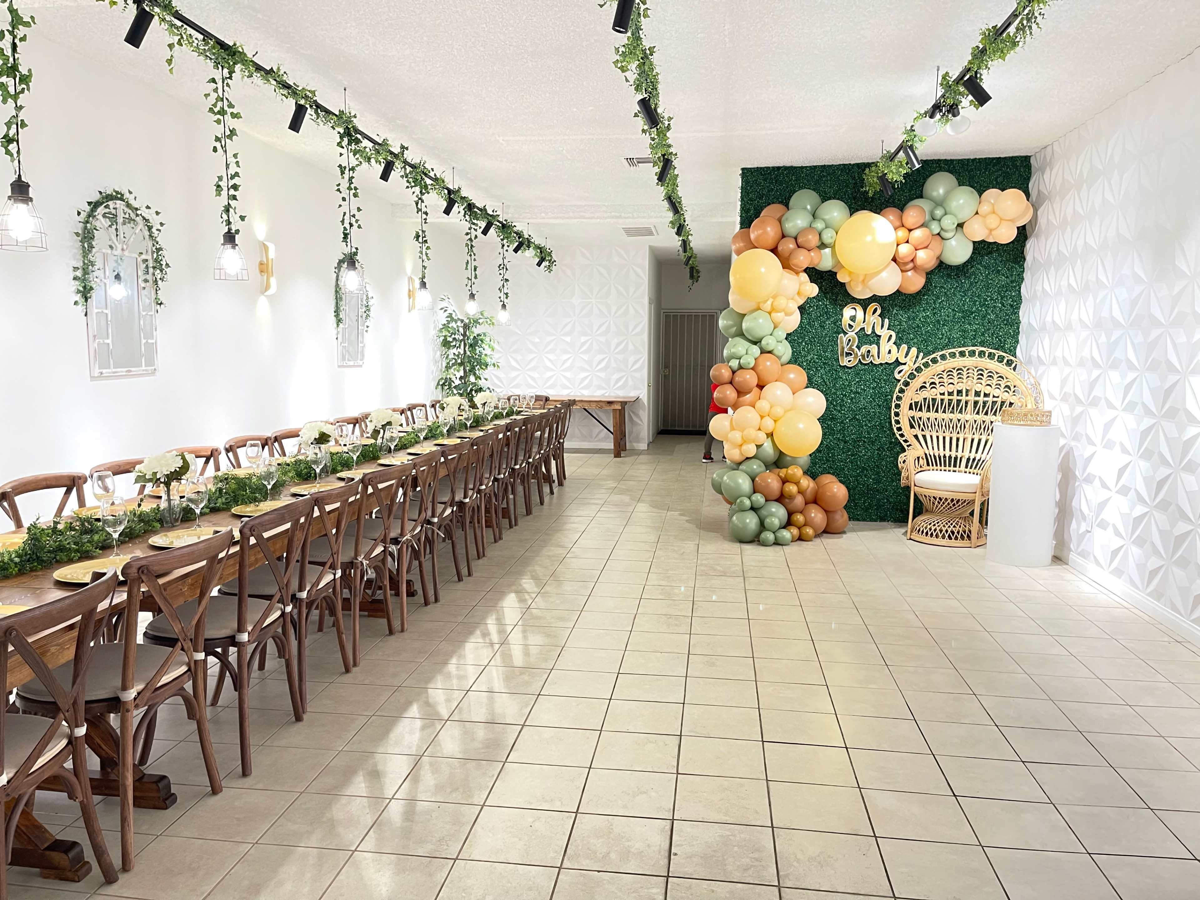 A spacious room is decorated with greenery and balloons, featuring a long wooden table and a cozy seating area against a textured wall.