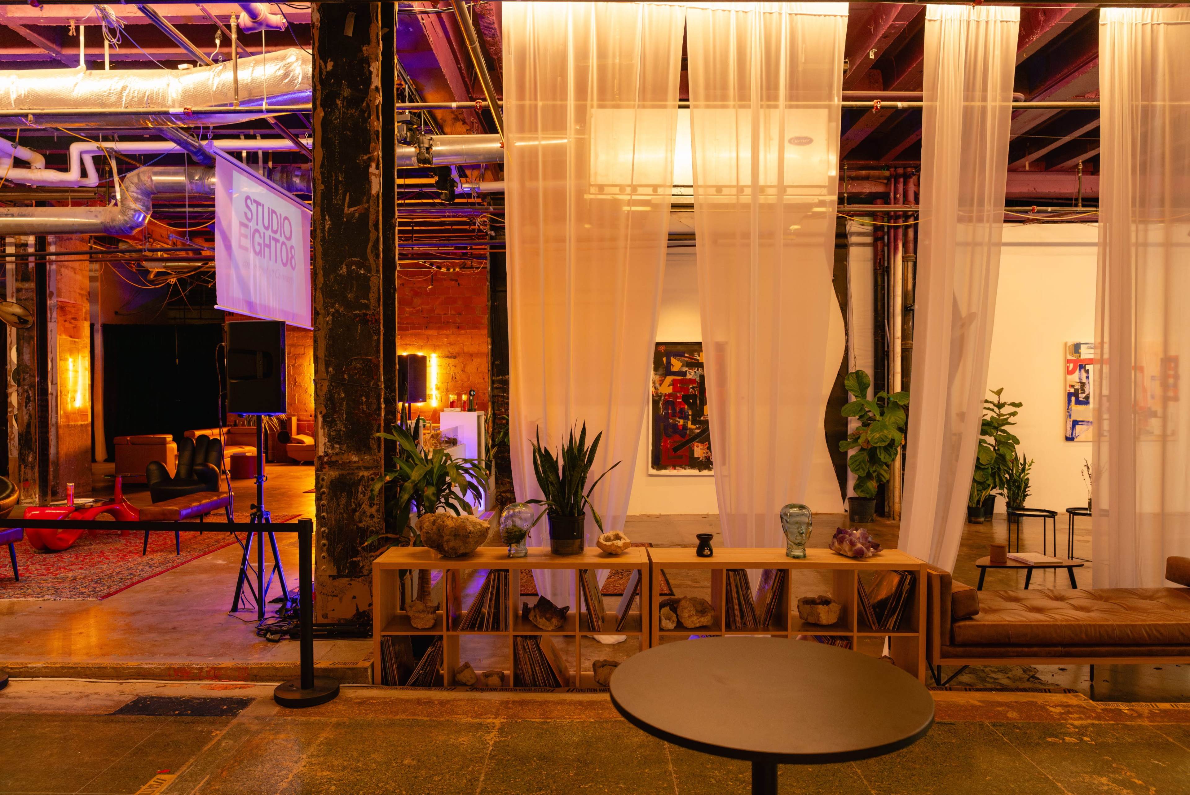 The image shows an art studio space with curtains dividing areas, a wooden bench displaying various items, and soft lighting illuminating the room.