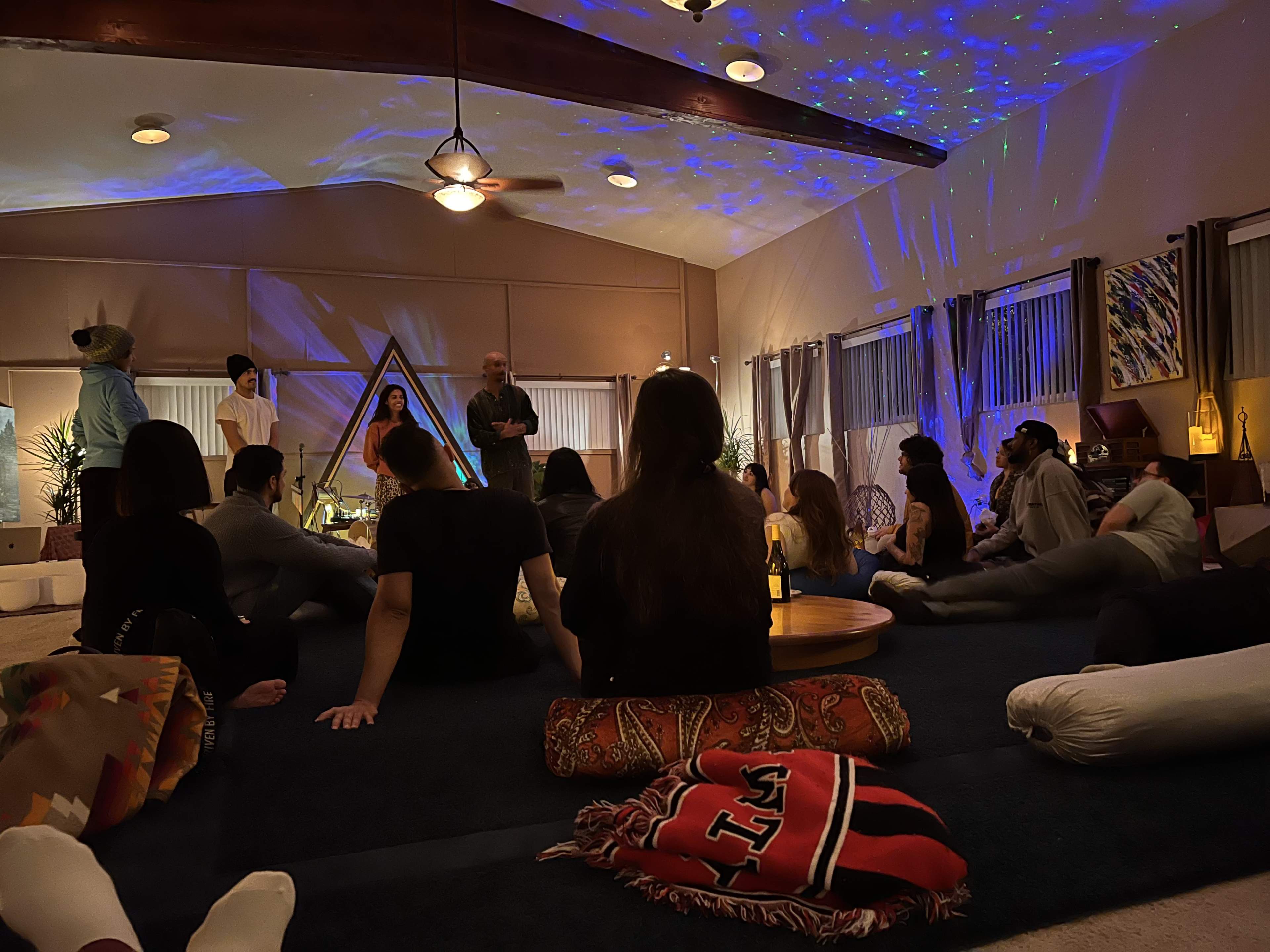 A group of people is seated on a carpeted floor in a dimly lit room, engaging in conversation while a light projector creates star patterns on the ceiling.