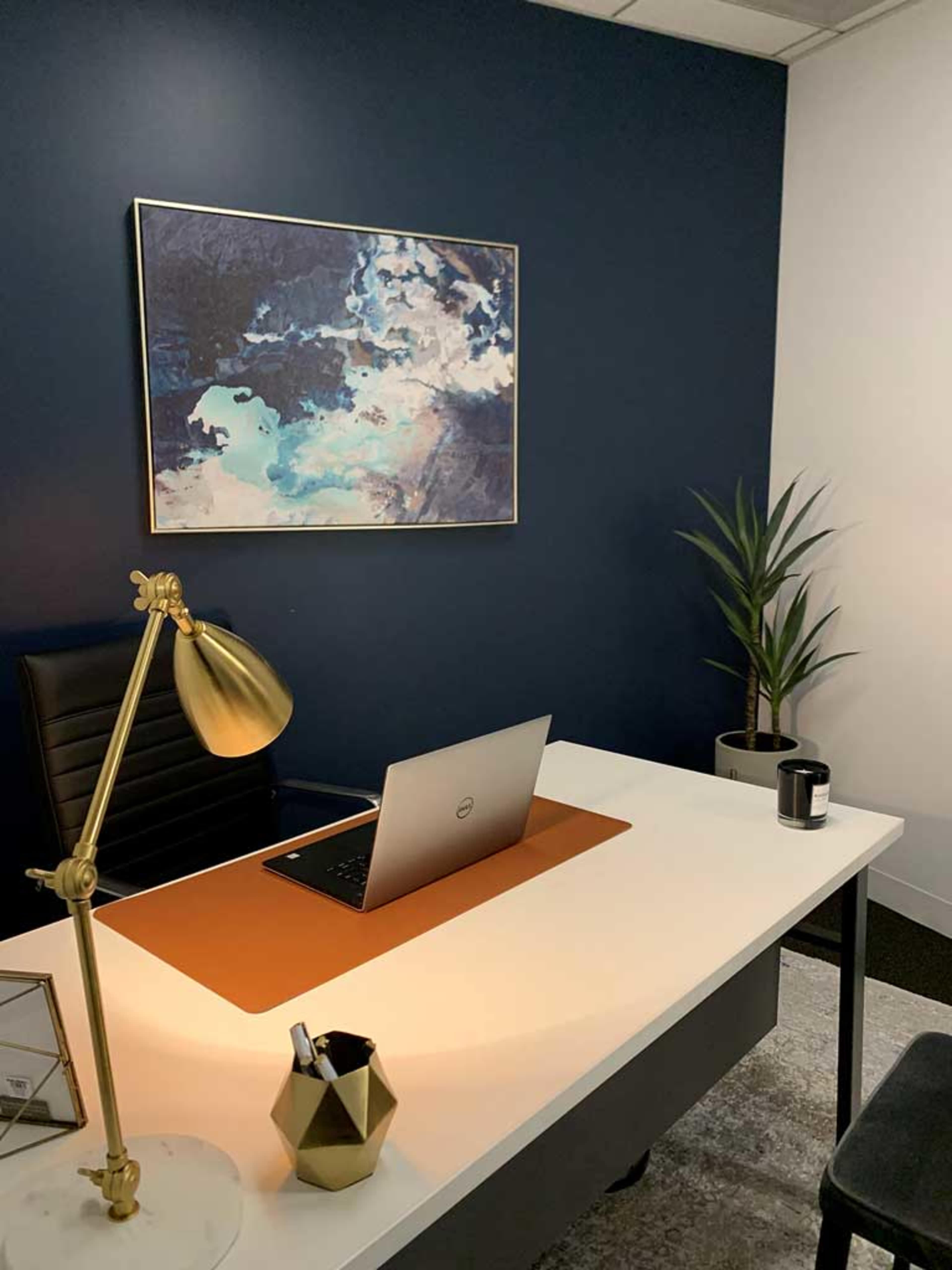 A modern office space features a white desk with a laptop, a golden lamp, a potted plant, and a blue accent wall adorned with abstract art.