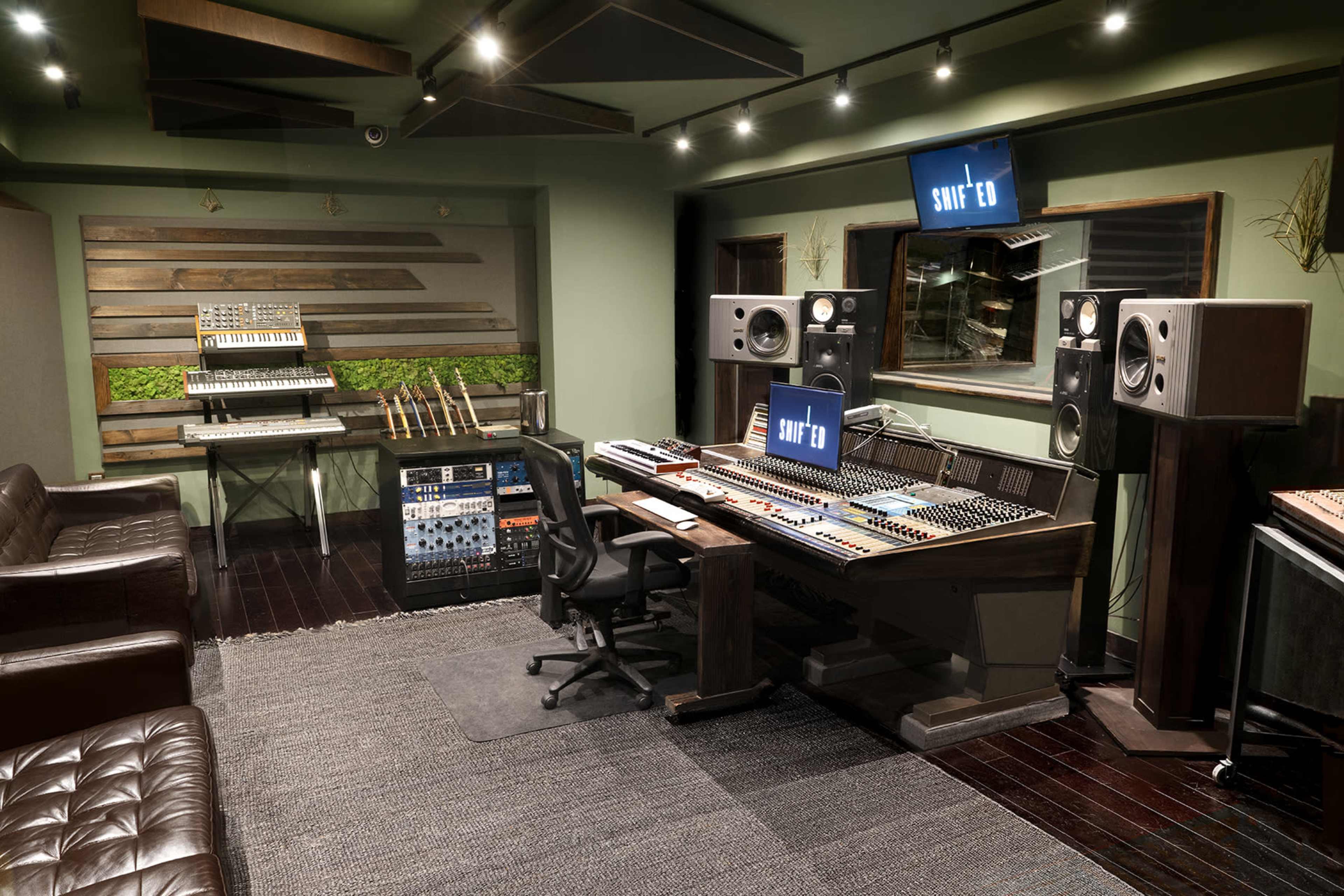 The image shows a recording studio featuring a large mixing console, audio equipment racks, and various musical instruments, with green walls and ambient lighting.