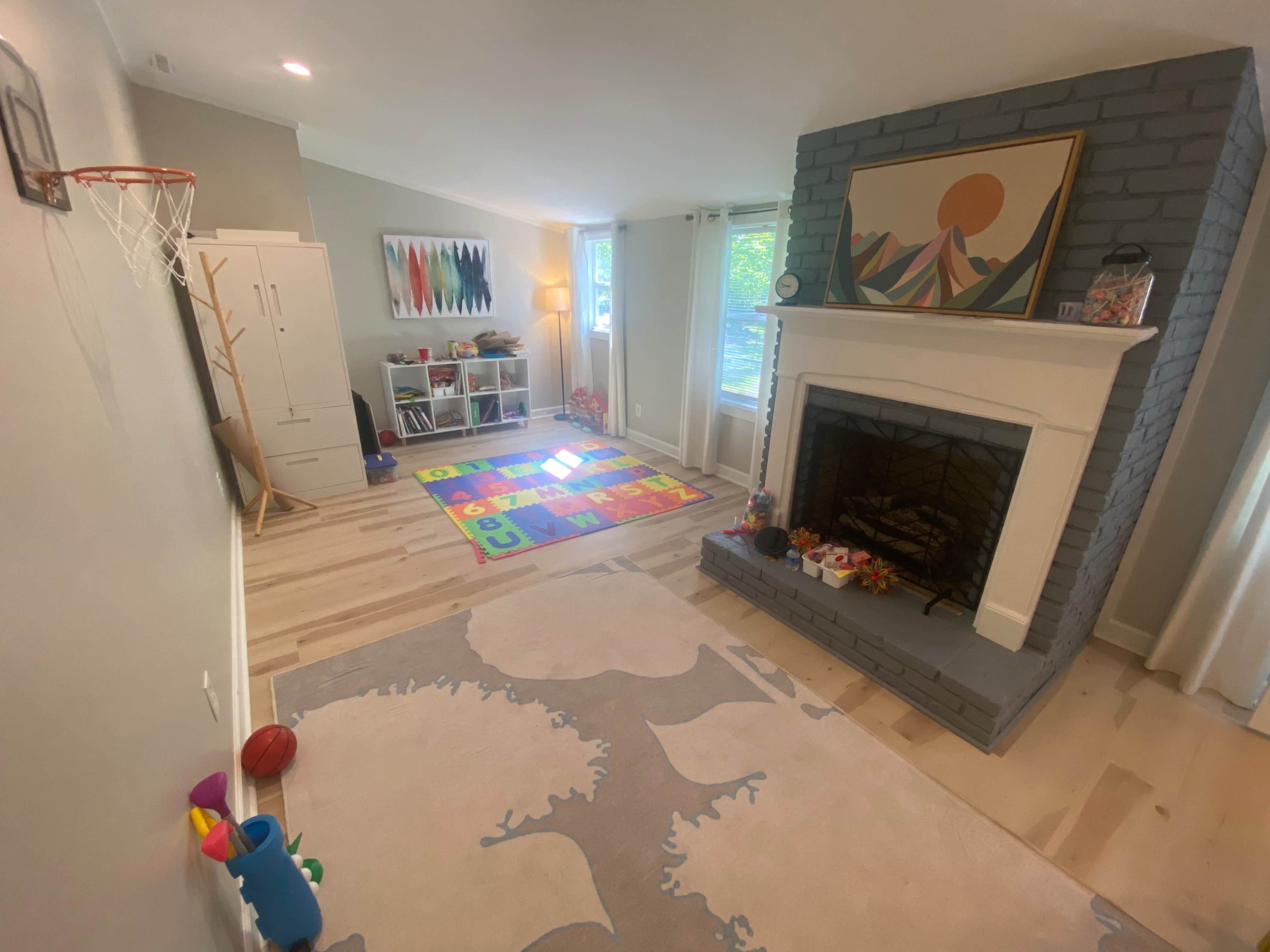 A playroom features a colorful rug with letters, a basketball hoop, a fireplace with a gray brick facade, and a window allowing natural light.