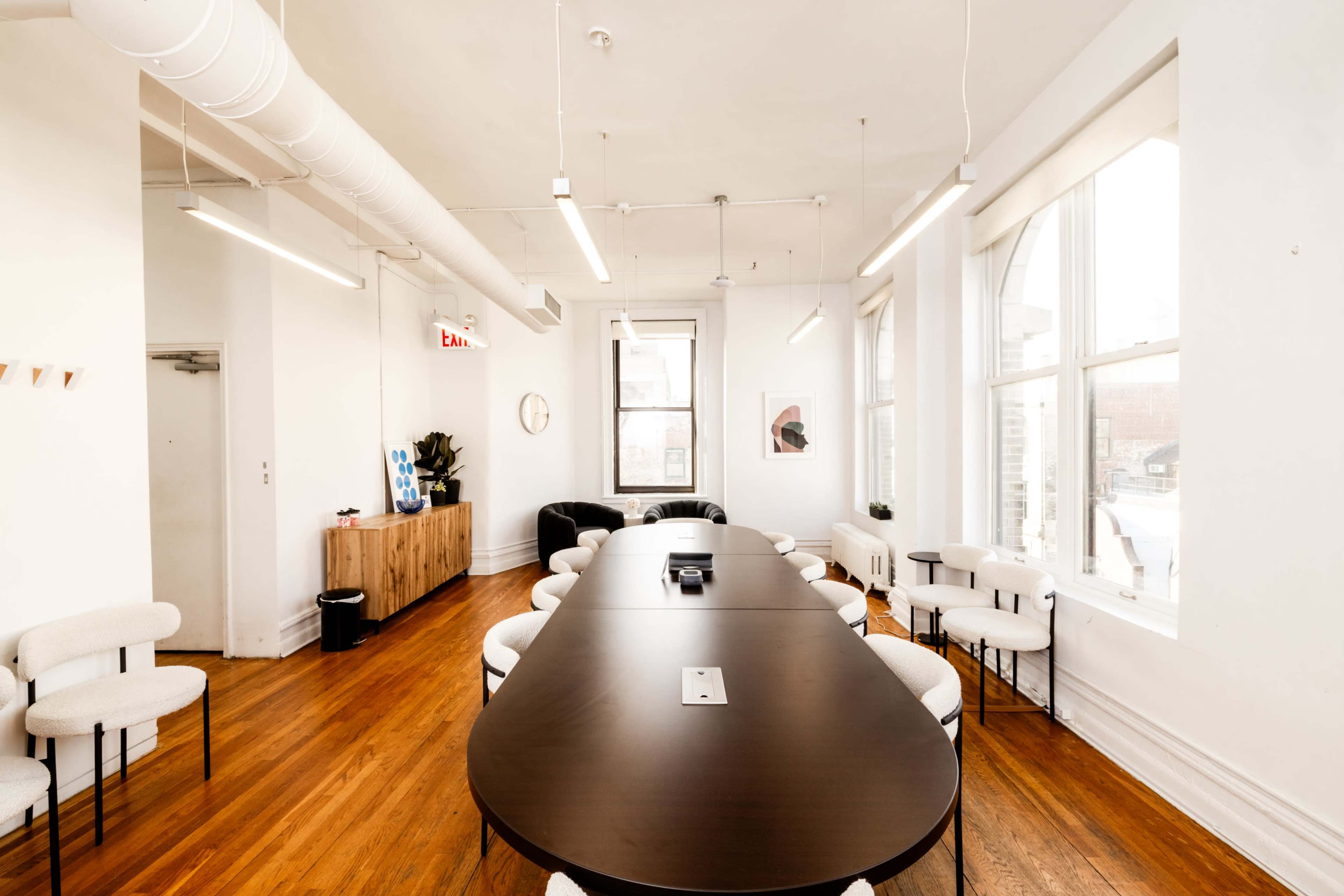 The image shows a bright conference room featuring a long, dark wooden table surrounded by white chairs, with large windows providing ample natural light.
