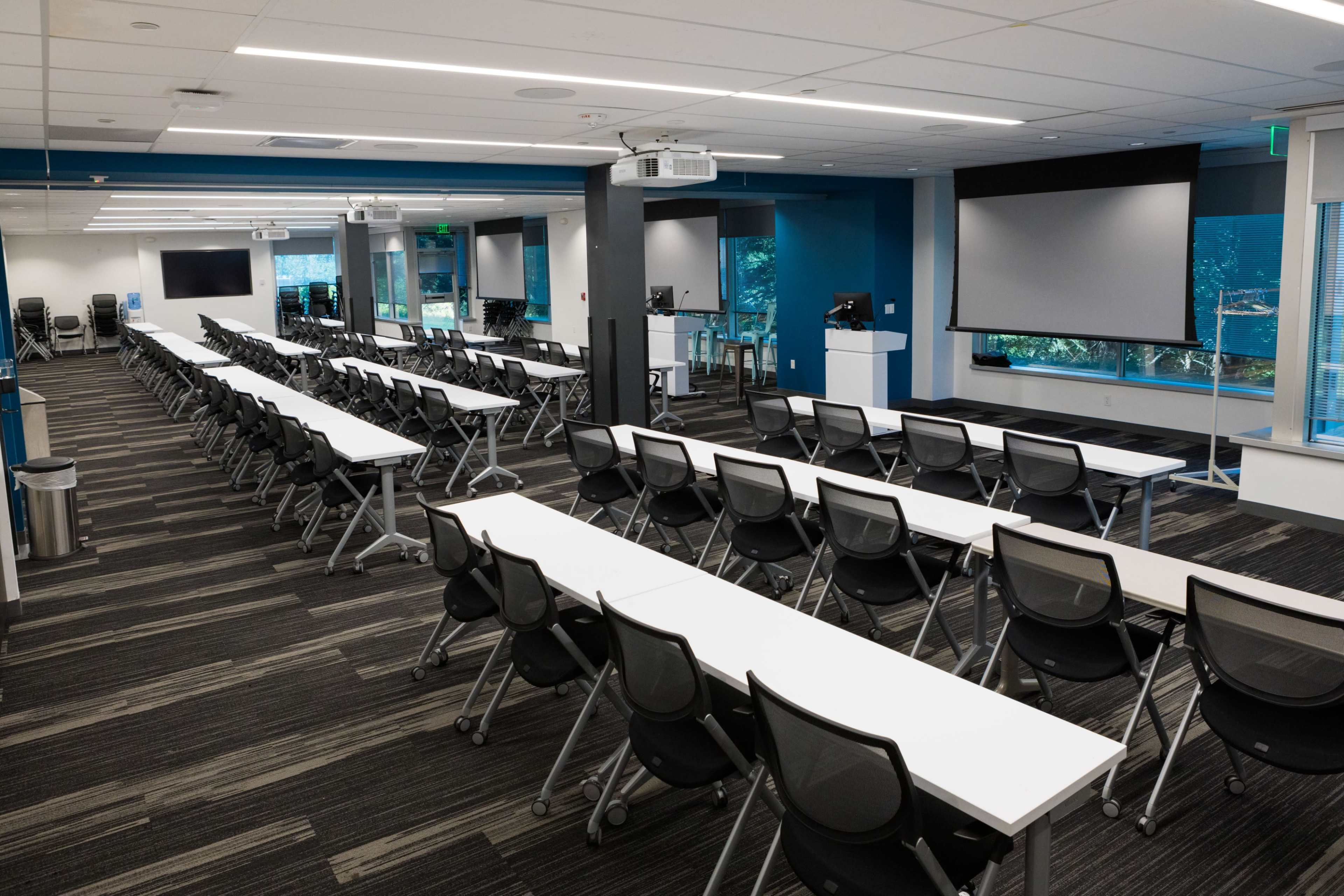 The image shows a modern classroom setup with rows of white tables and black chairs arranged for a training session or meeting, featuring large screens and windows that provide natural light.