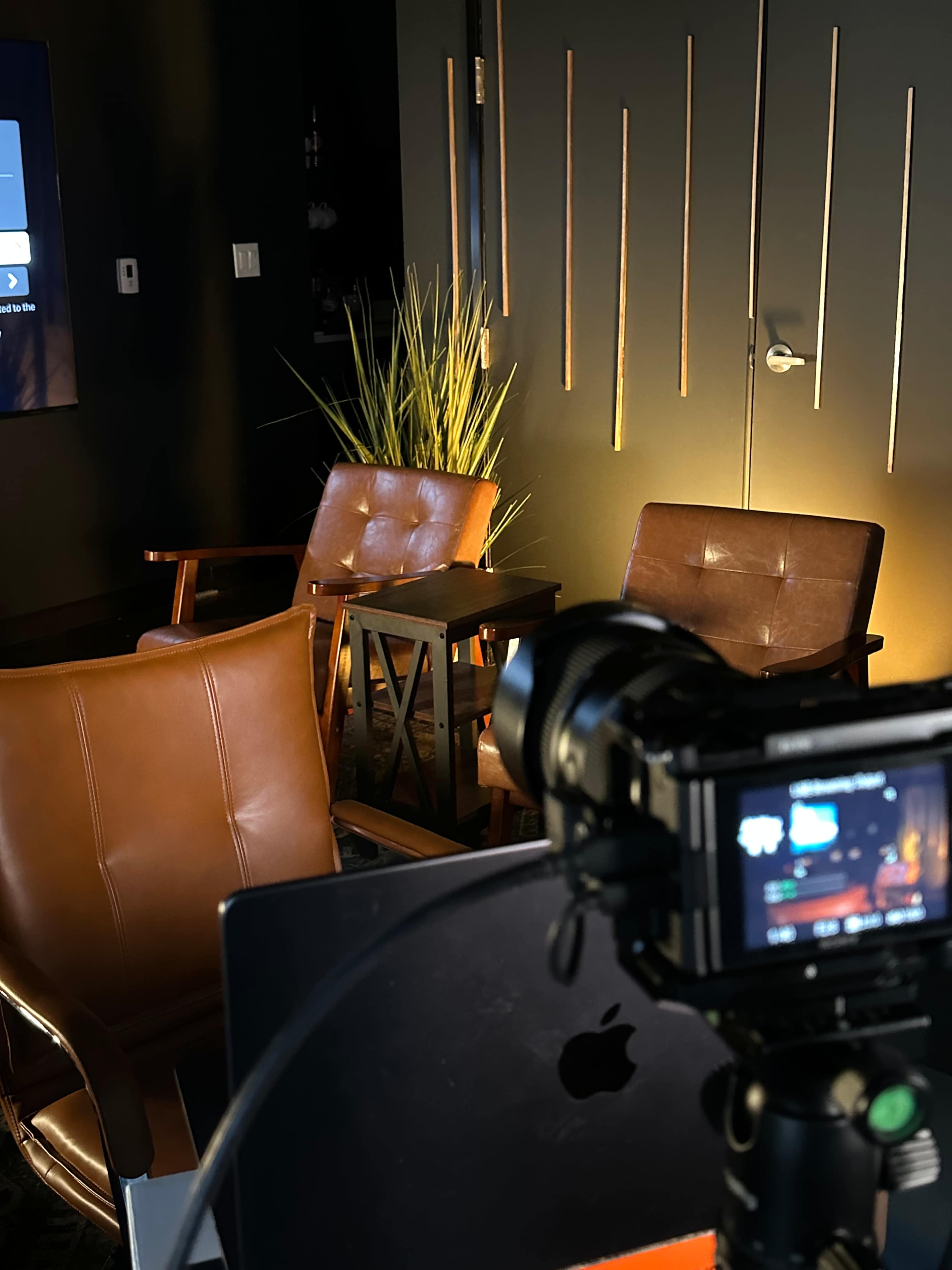 The image shows a dimly lit room featuring two brown leather chairs, a small table, and a camera pointed towards a television screen.