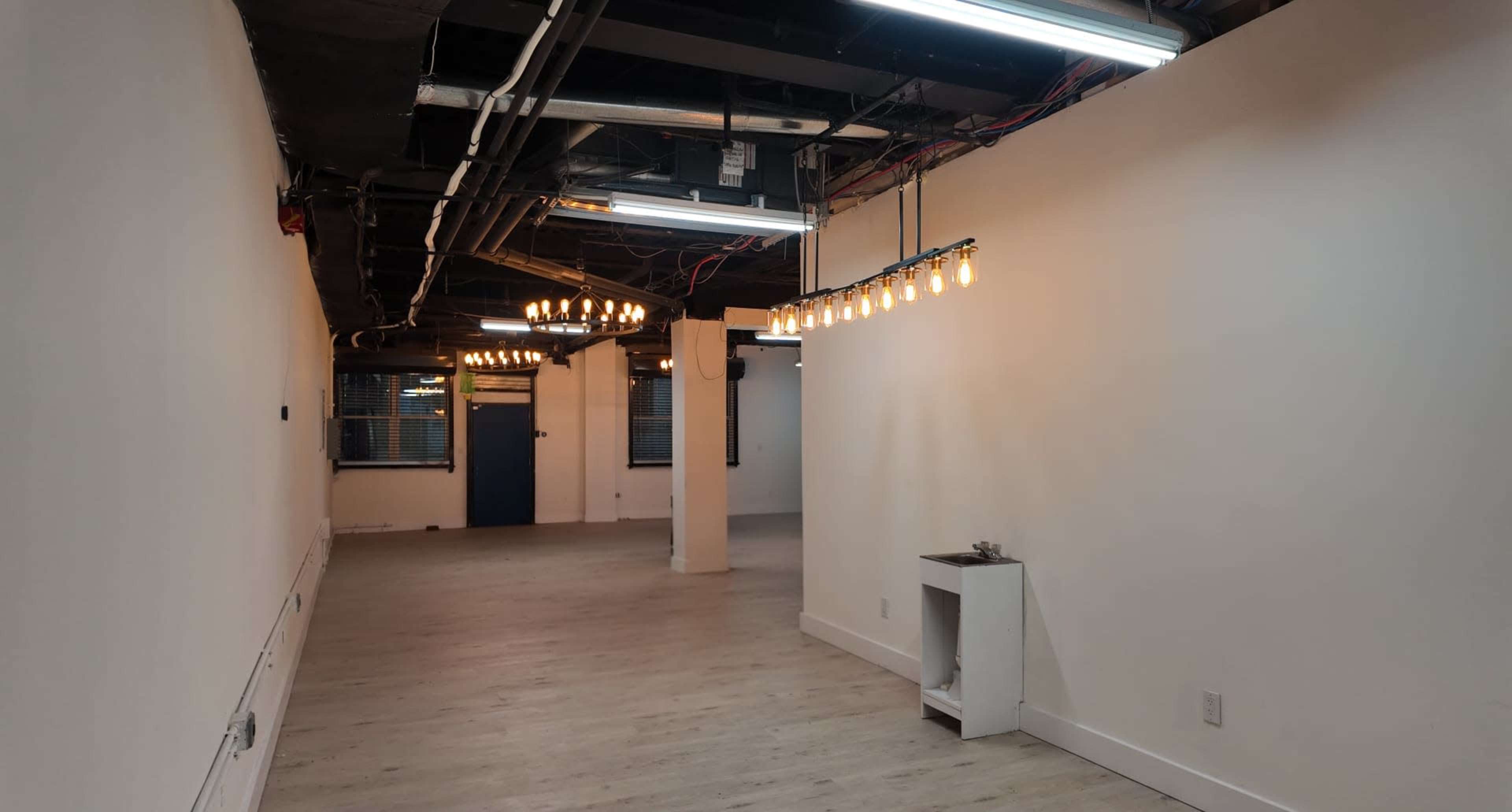 The image shows a spacious, well-lit interior of a room with exposed ceilings, a row of hanging light bulbs, and a small fixture against one wall.