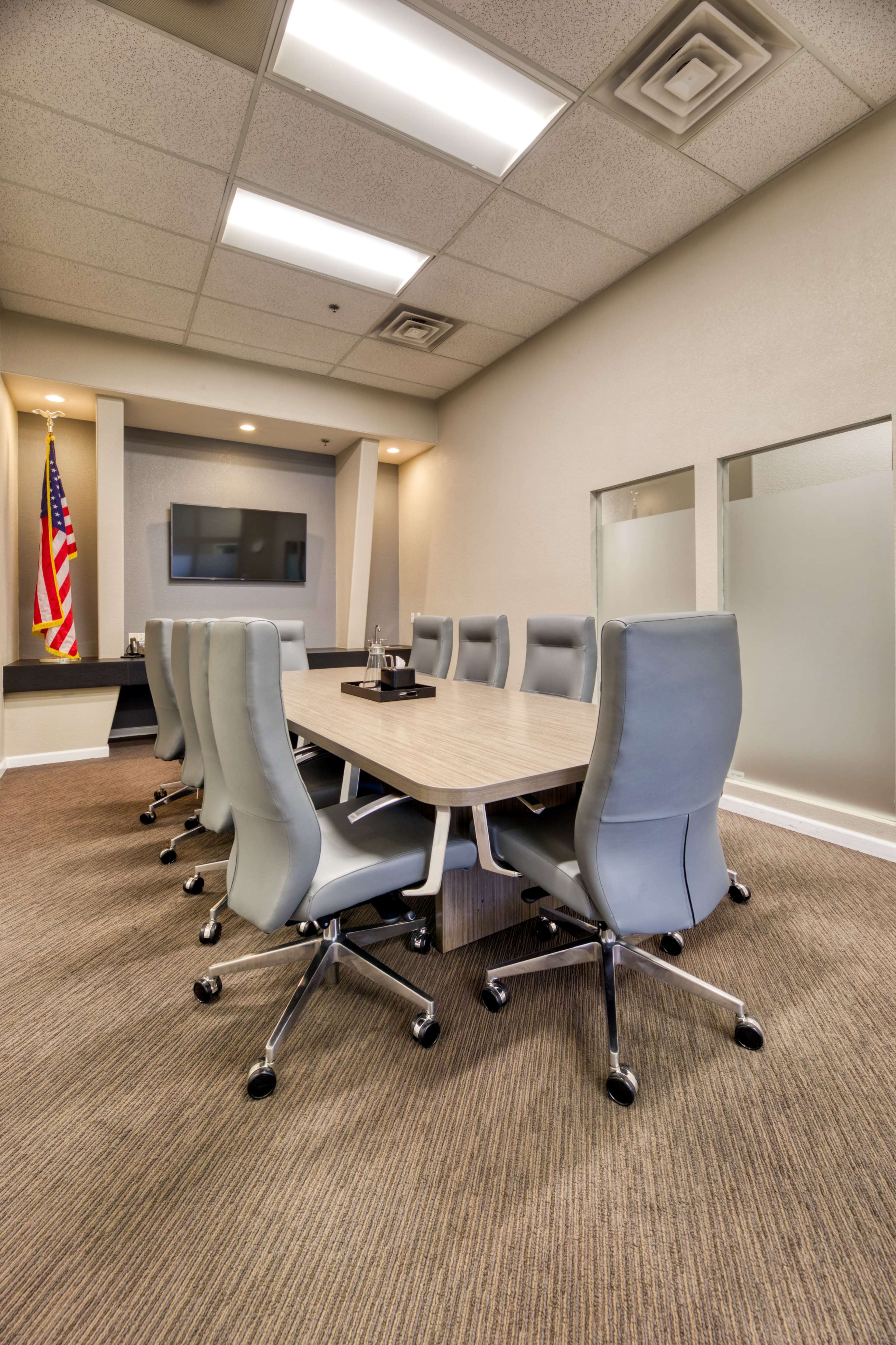 A modern conference room features a long wooden table surrounded by ten gray office chairs, with an American flag displayed in the corner.