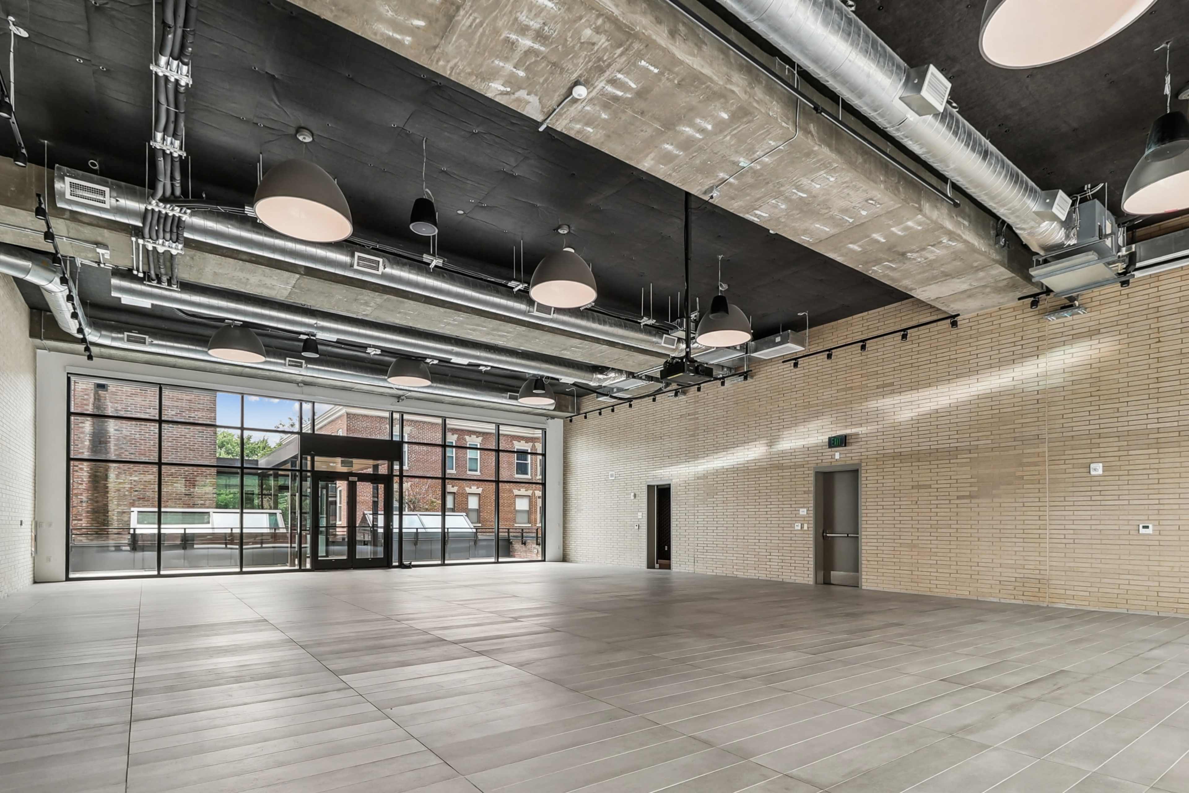 The image shows a spacious interior room with a polished tile floor, high ceilings, and large windows that allow natural light to enter.