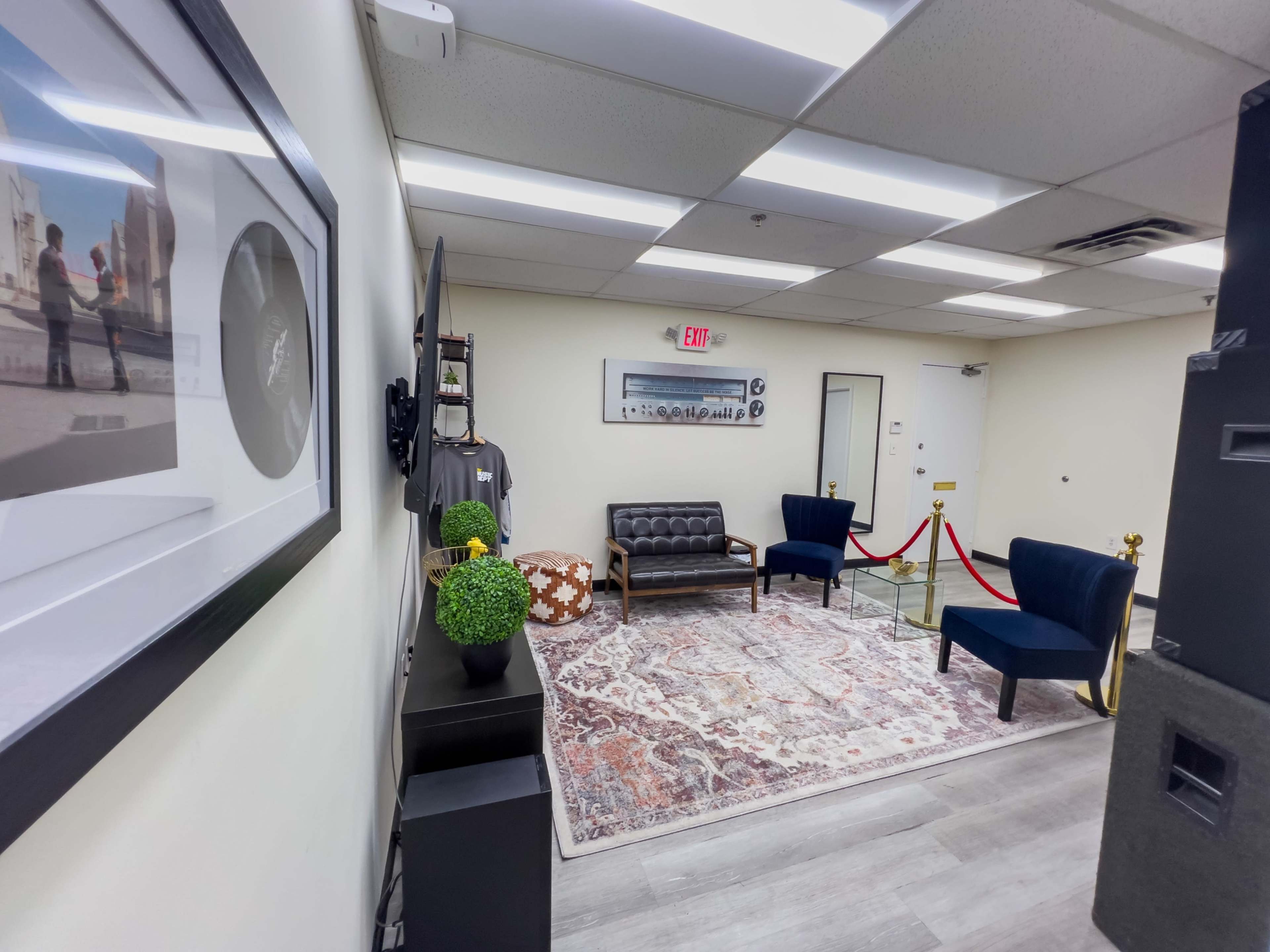 A small, modern lounge area with a black leather couch, two blue chairs, and decorative plants, surrounded by a rug and a barrier rope.