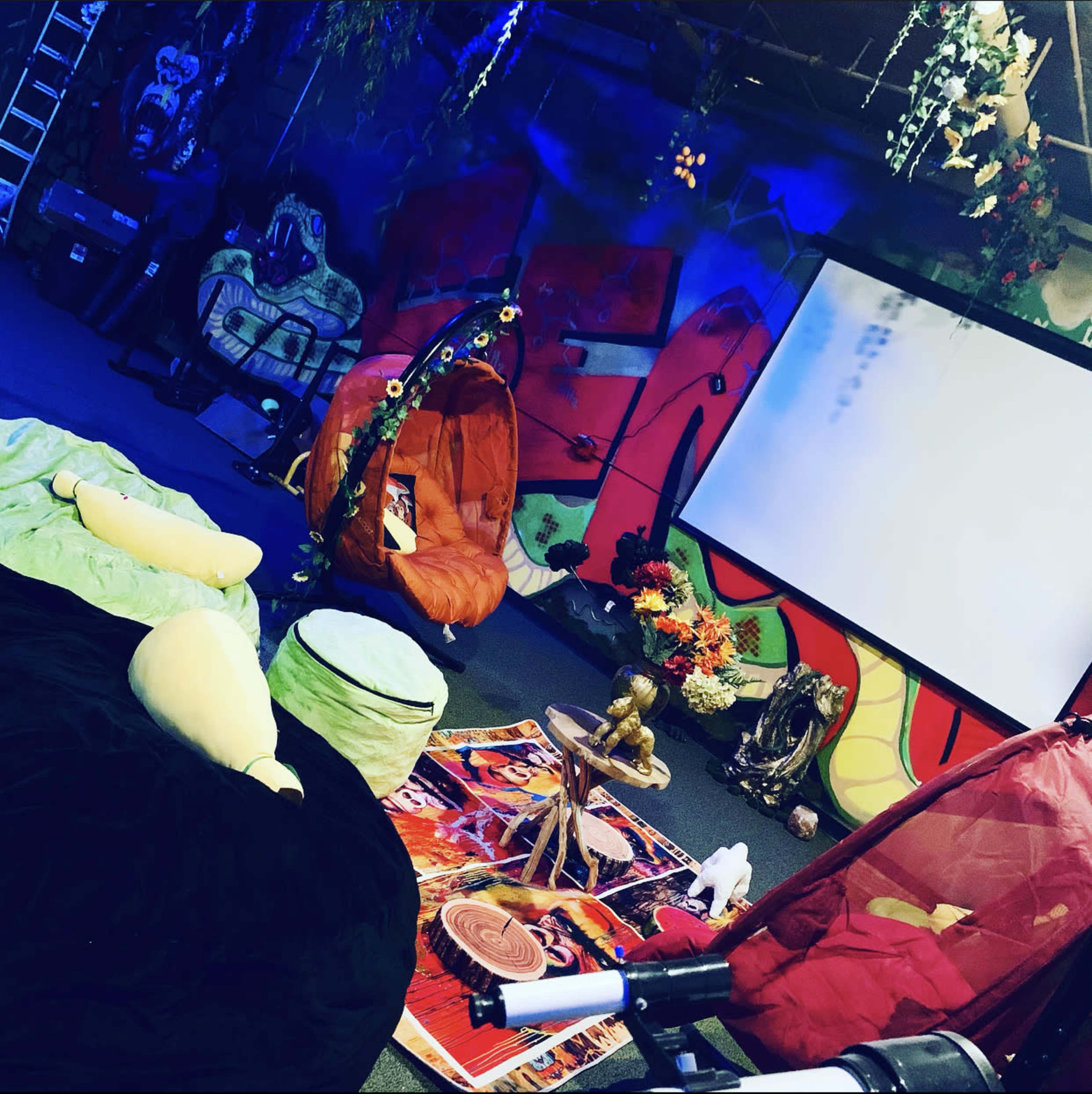 An eclectic room featuring bean bag chairs, a large screen, and colorful wall murals, with various decorative elements scattered throughout.