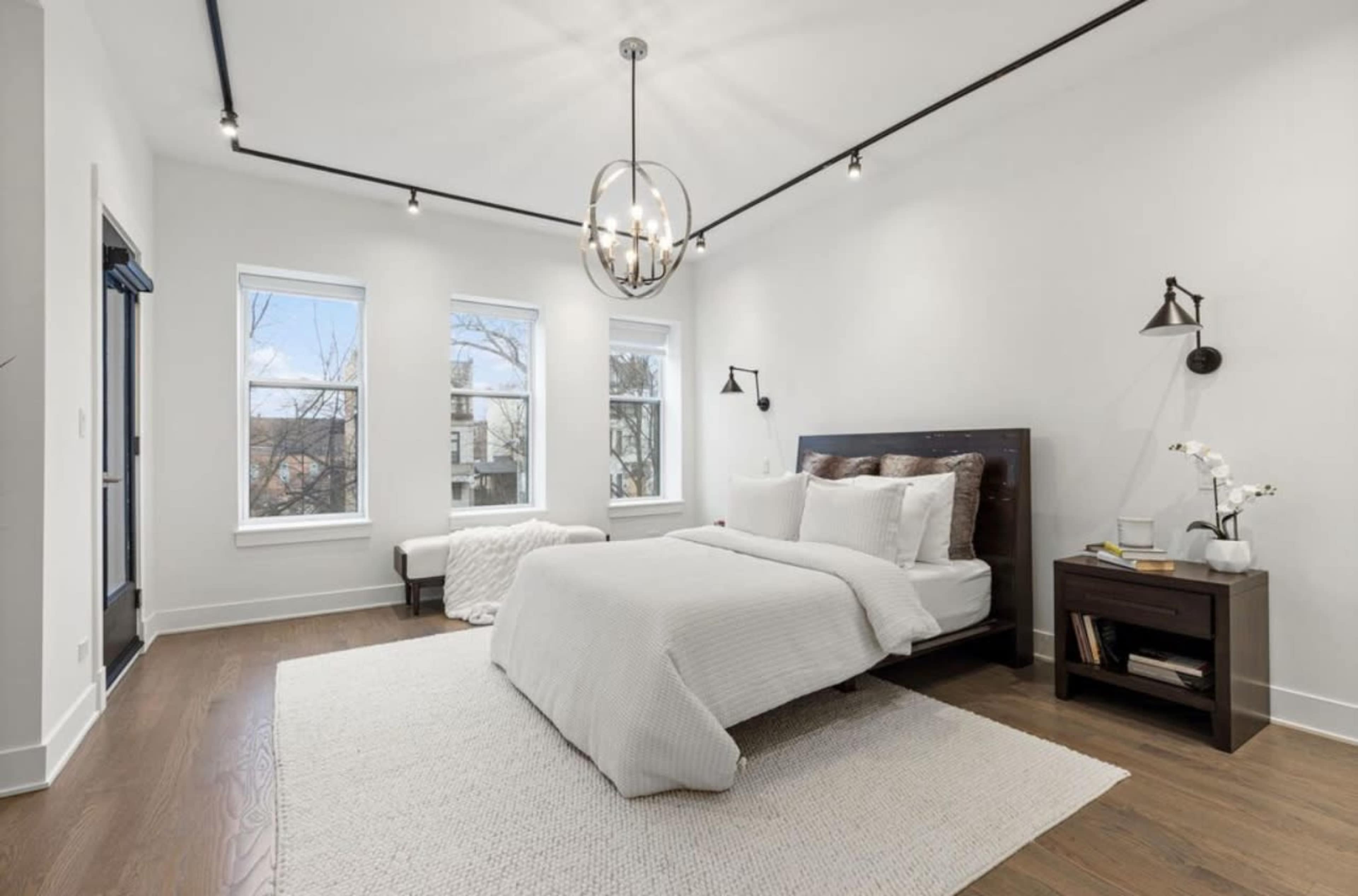 The image shows a bright bedroom with a bed, nightstand, and large windows allowing natural light to enter.