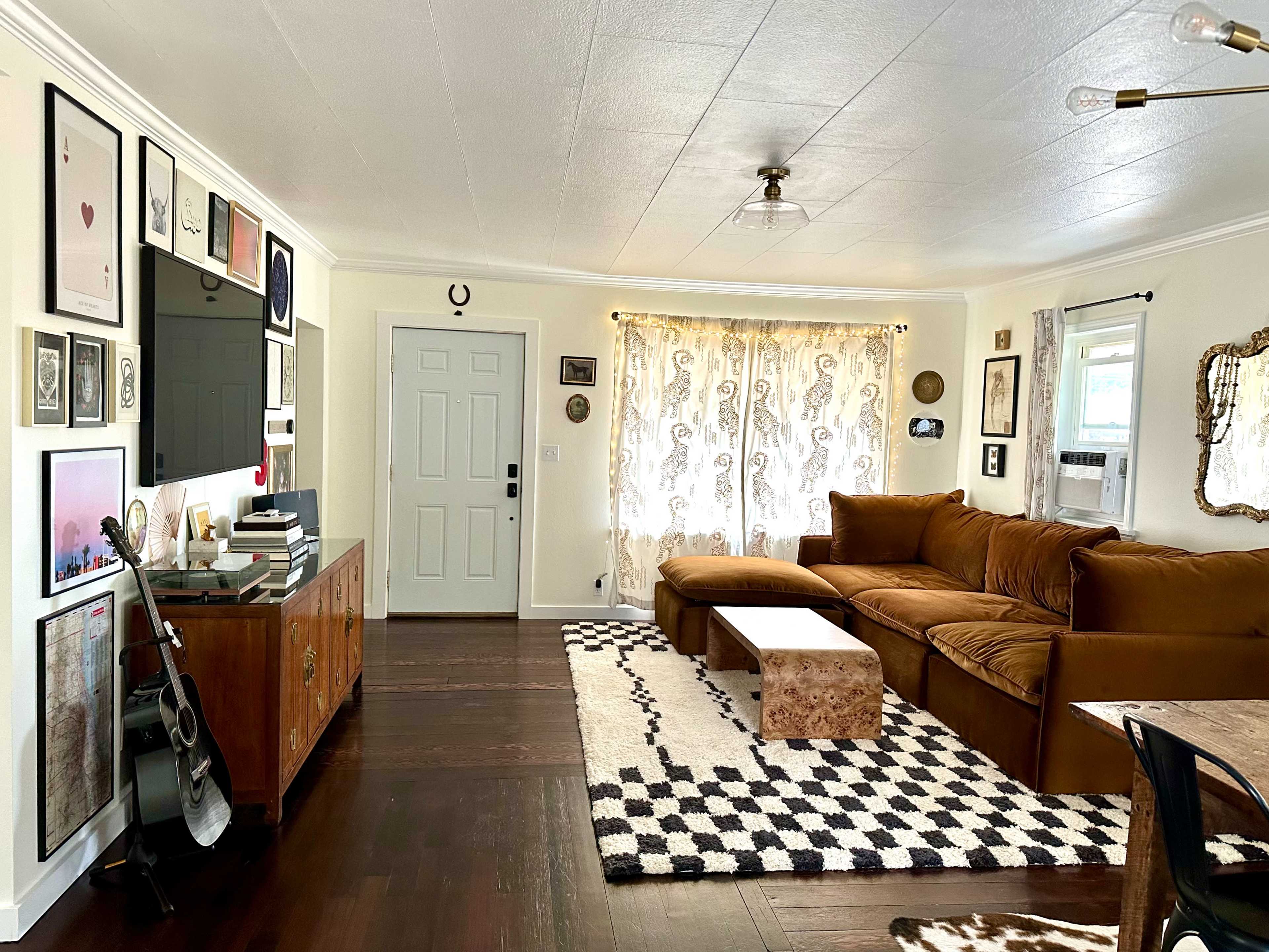 The image shows a cozy living room with a brown sectional sofa, patterned rug, wooden media console, and framed artwork on the walls.