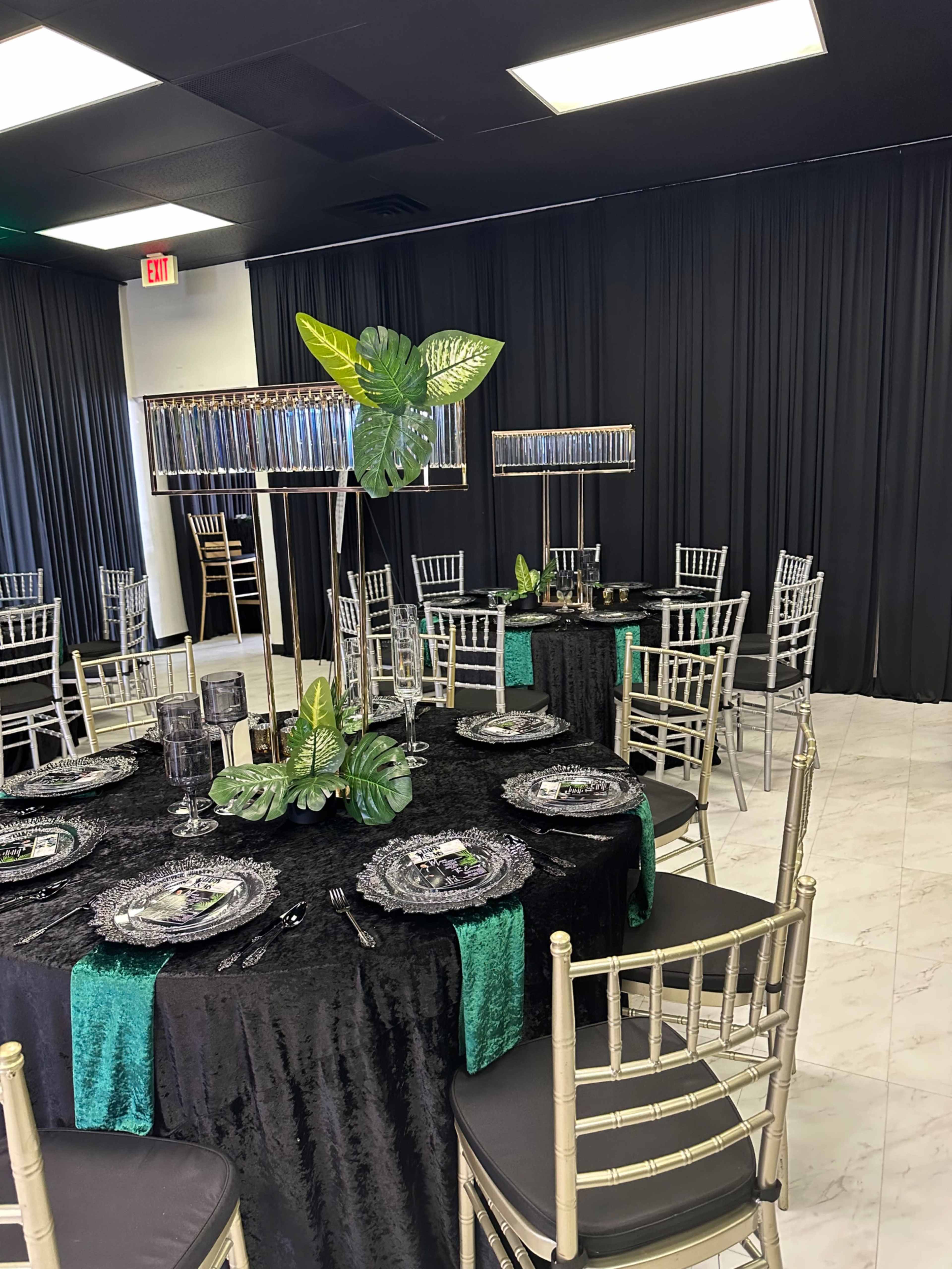 The image shows a dining area set up with elegantly arranged tables covered in black and green tablecloths, adorned with decorative centerpieces and surrounded by gold chiavari chairs.