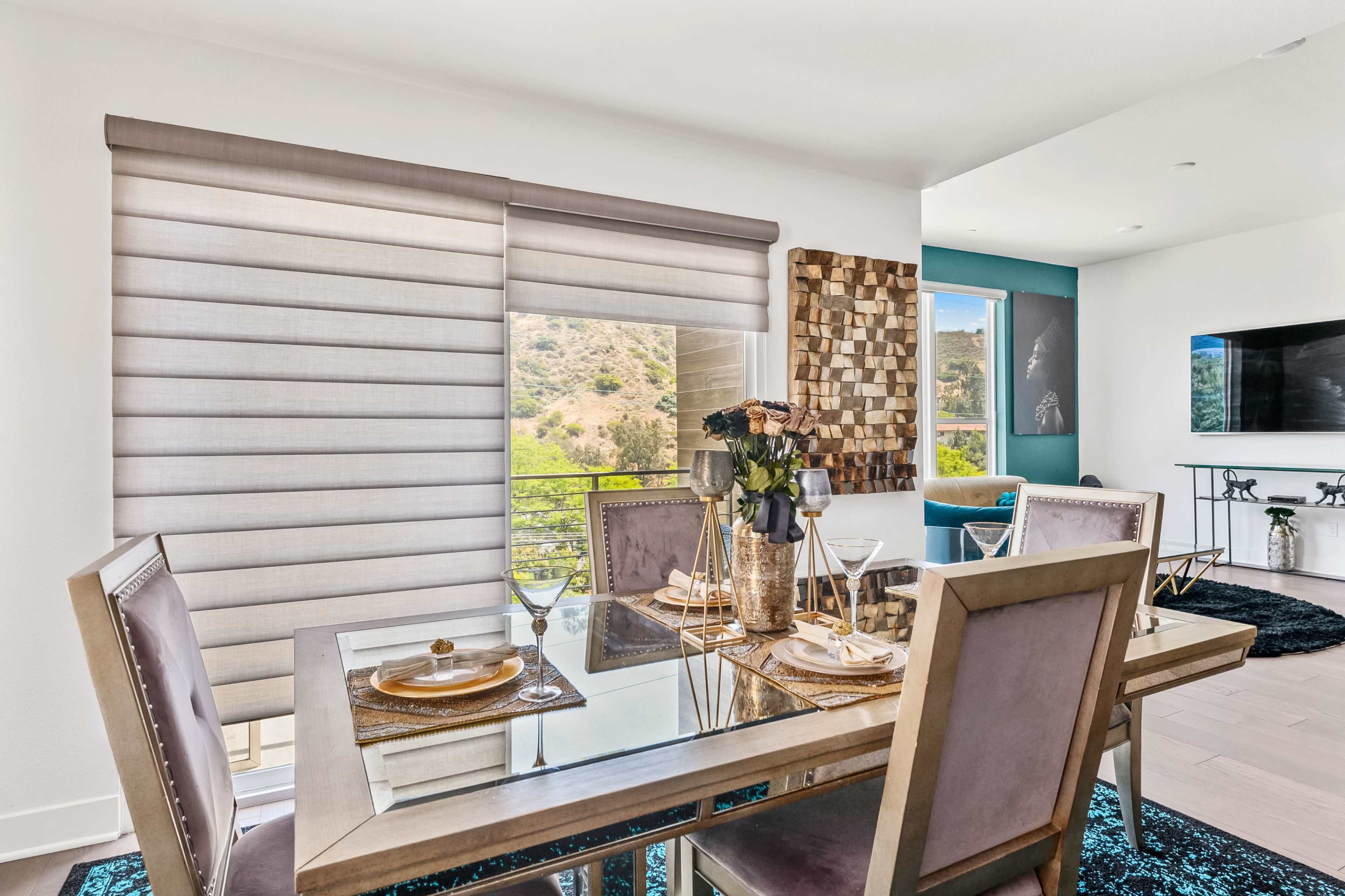 A modern dining area features a glass table set with plates and glasses, surrounded by plush chairs, near large windows with layered shades overlooking a green landscape.