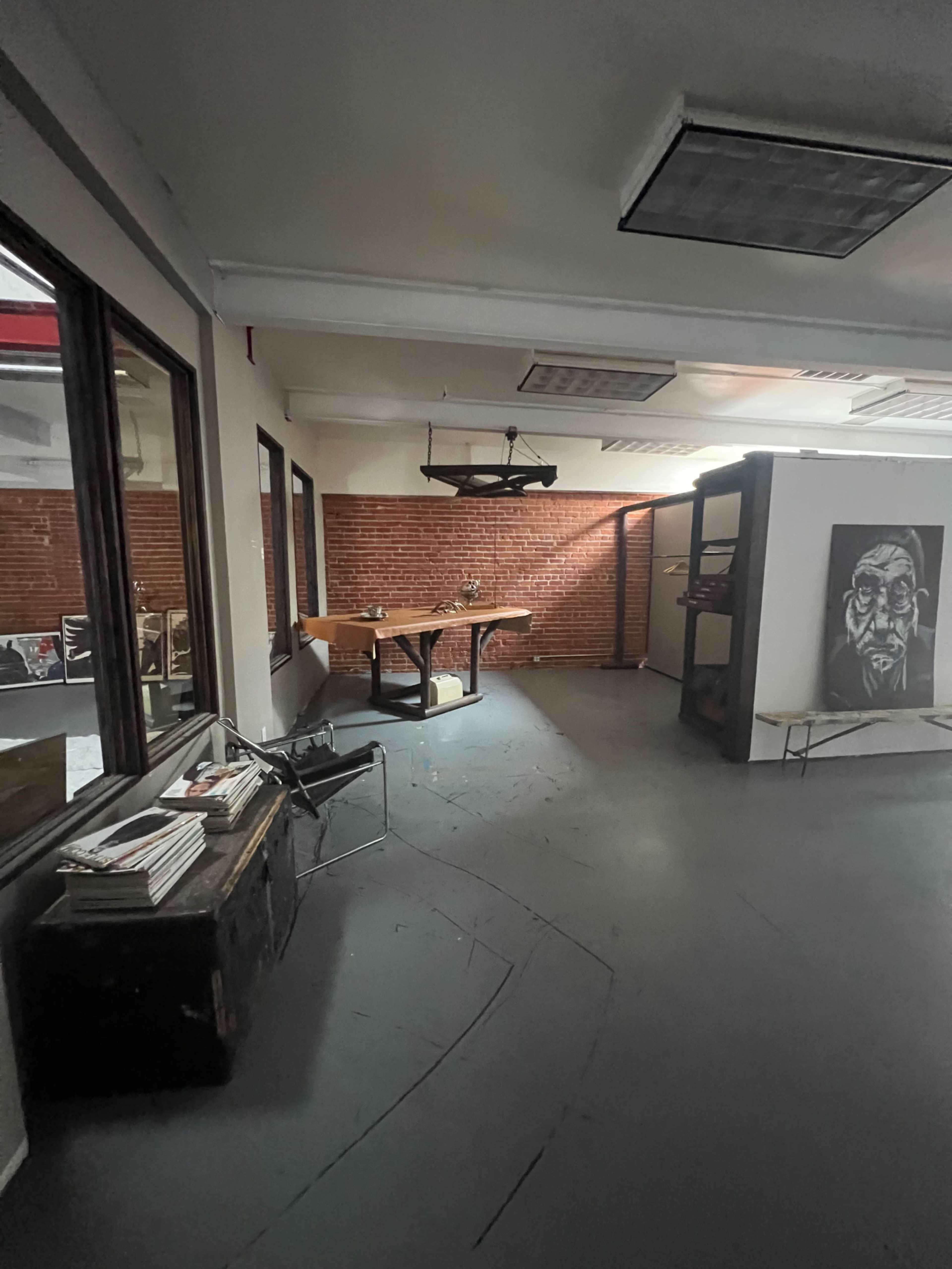 The image shows an interior space with exposed brick walls, a large table in the center, and various items arranged around a sparse, open area.