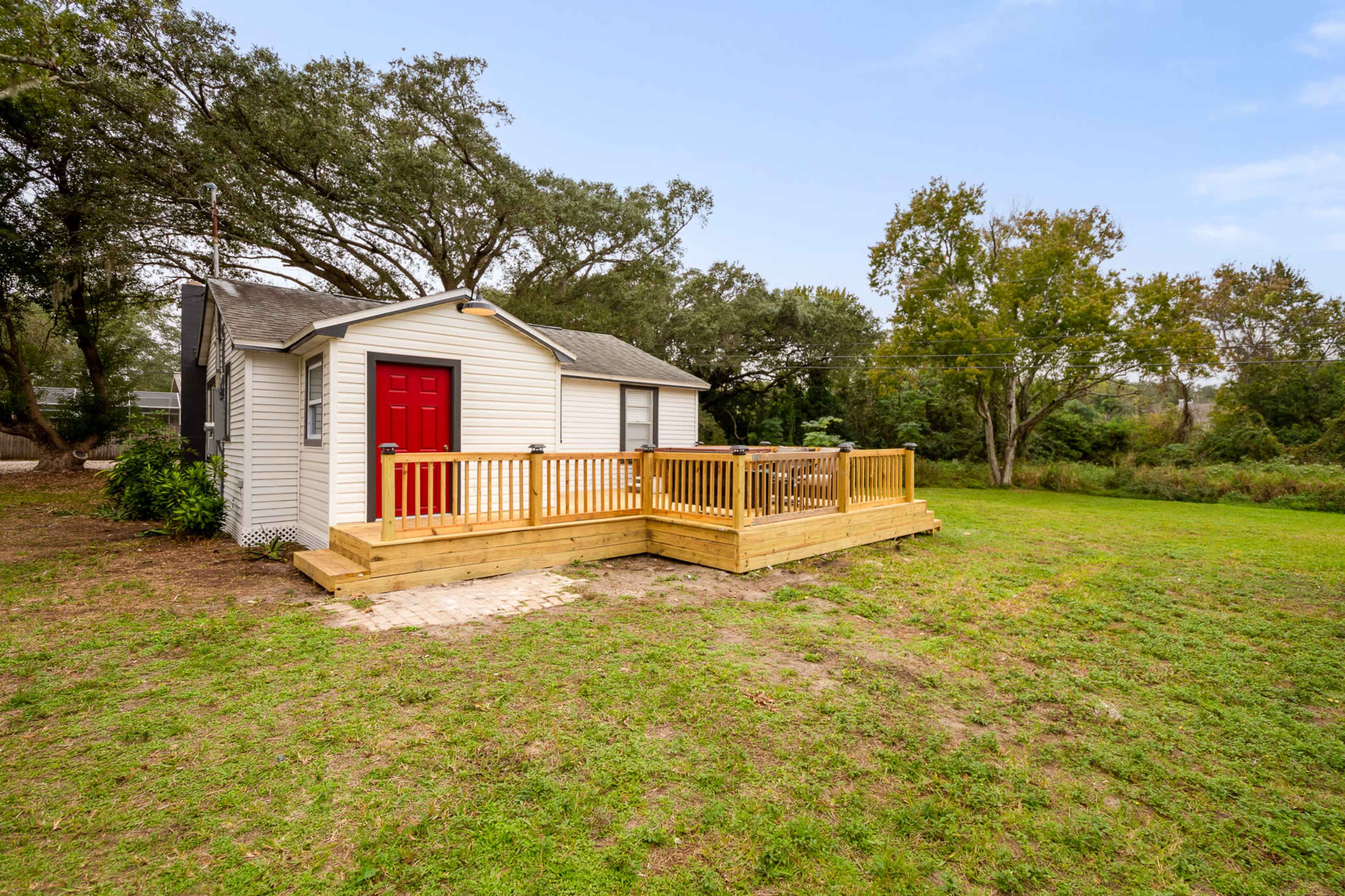 A small white house with a red door and a wooden deck in an open grassy area surrounded by trees.