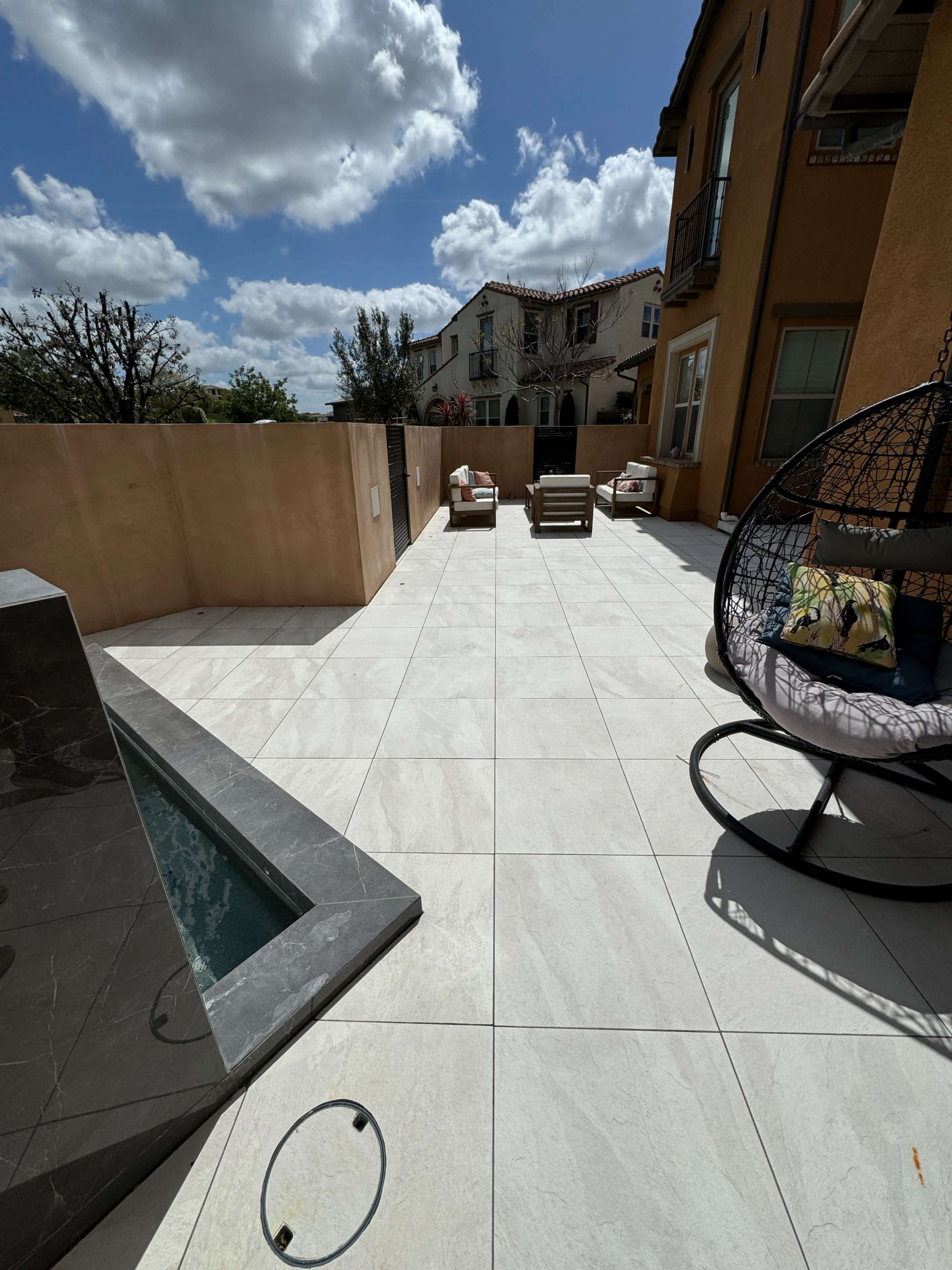 A spacious outdoor patio with a small pool, lounge chairs, and a hanging chair, surrounded by a low wall and adjacent buildings.