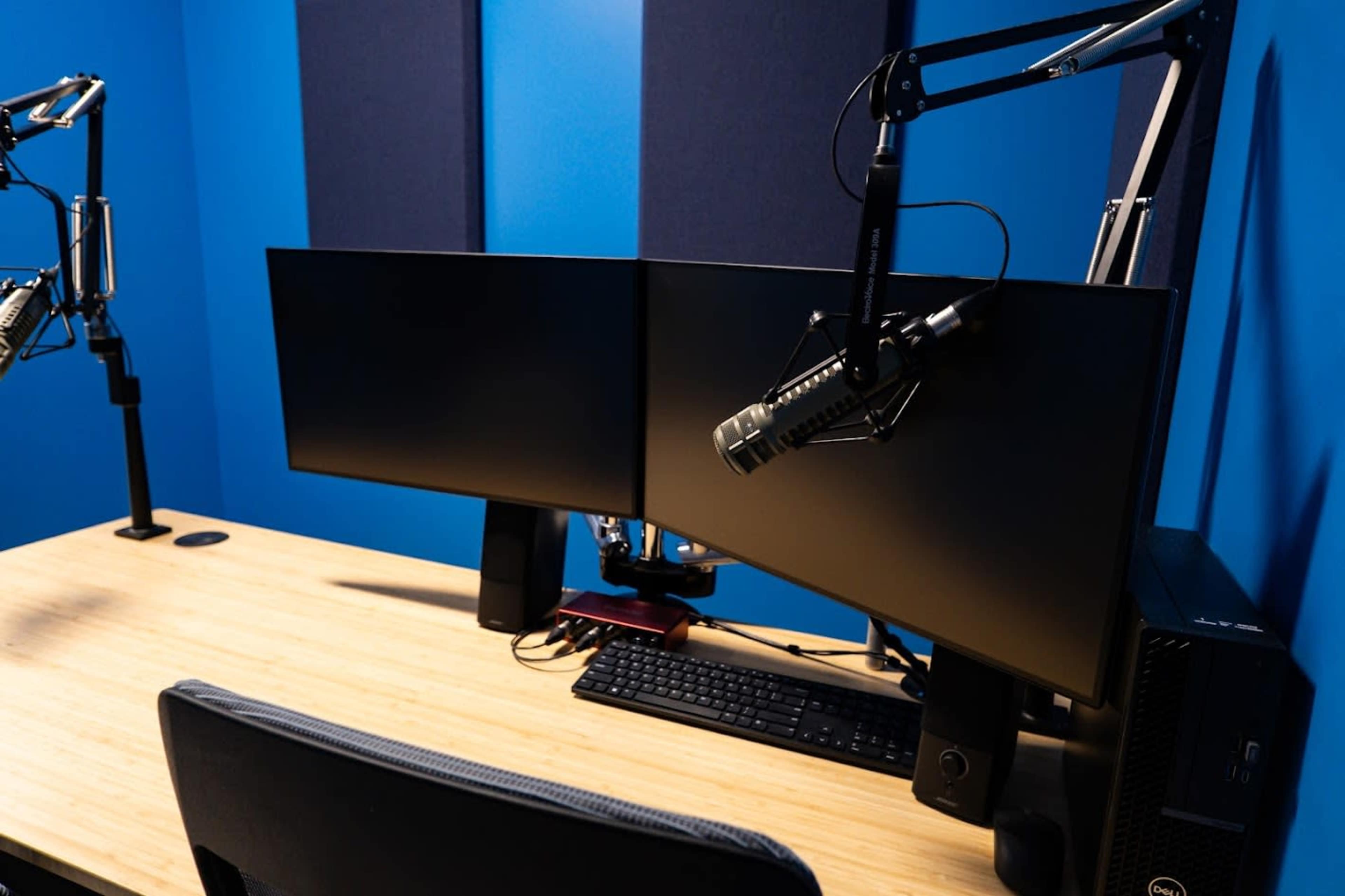 The image shows a modern workspace featuring a wooden desk with two curved computer monitors, a microphone mounted on an adjustable arm, and a keyboard.