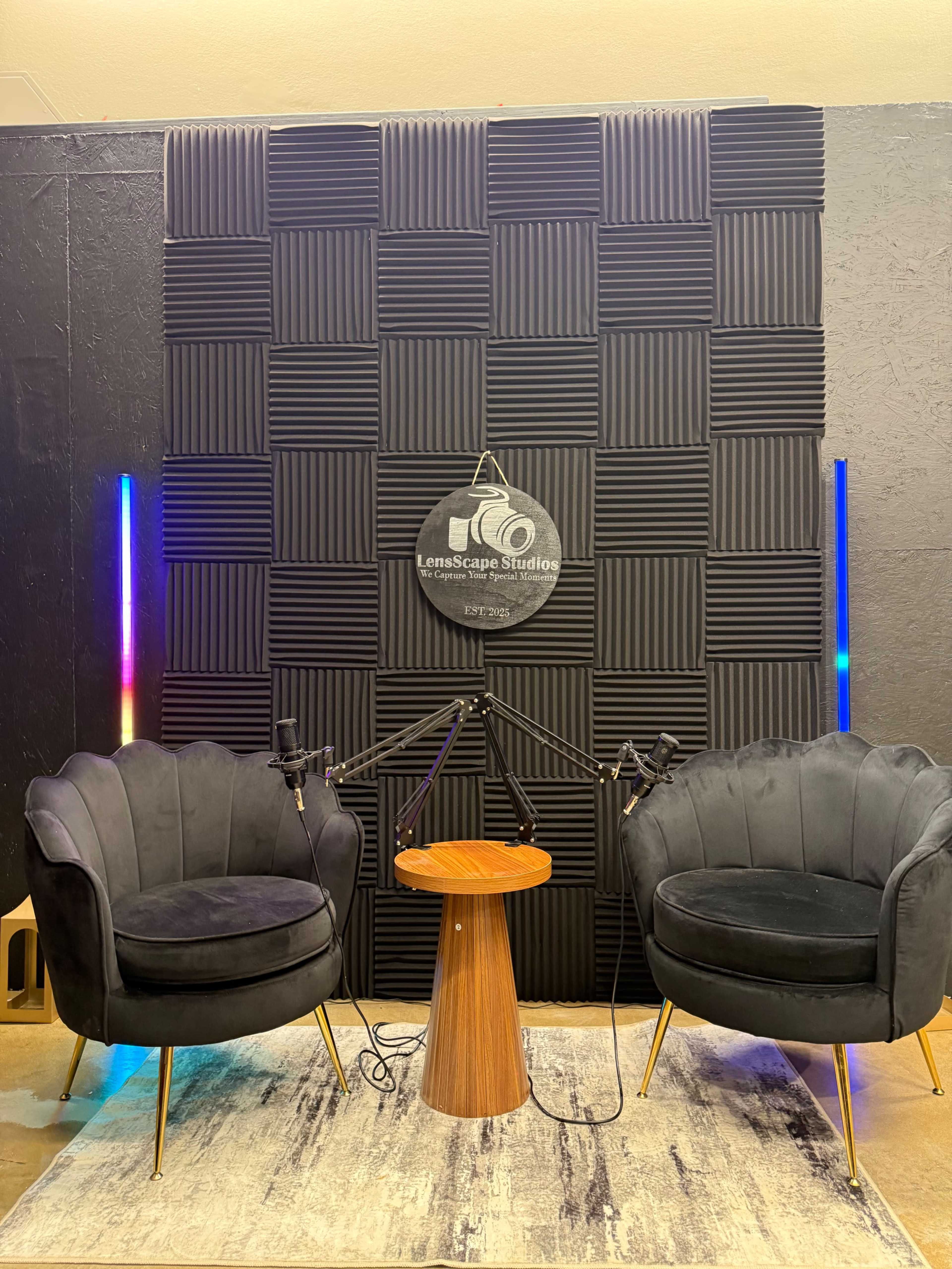 The image shows a podcast recording setup featuring two black velvet armchairs, a round wooden table, and microphones, all arranged in front of a soundproofed wall.