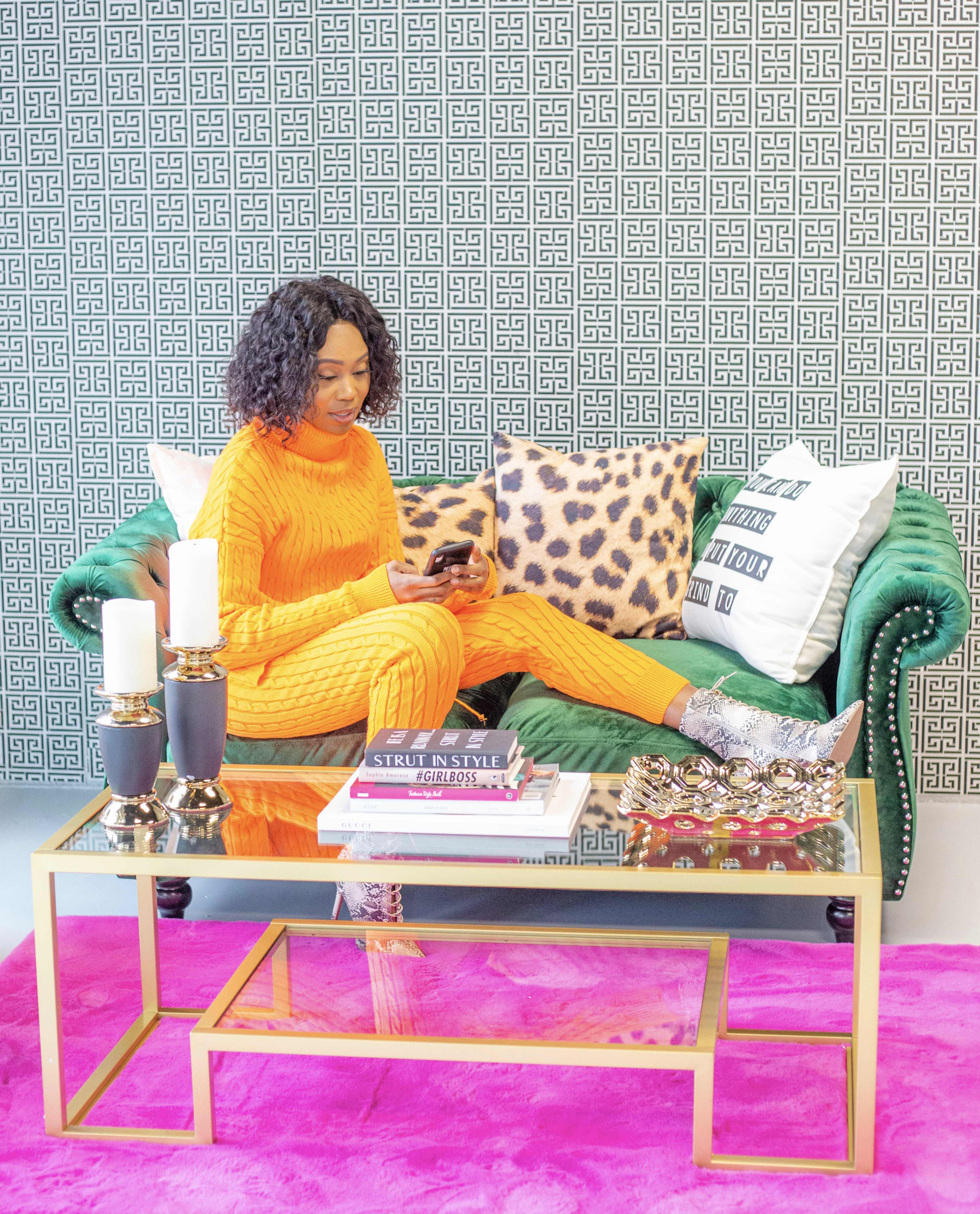 A woman in an orange outfit sits on a green couch, using her phone, surrounded by decorative pillows and a glass coffee table.