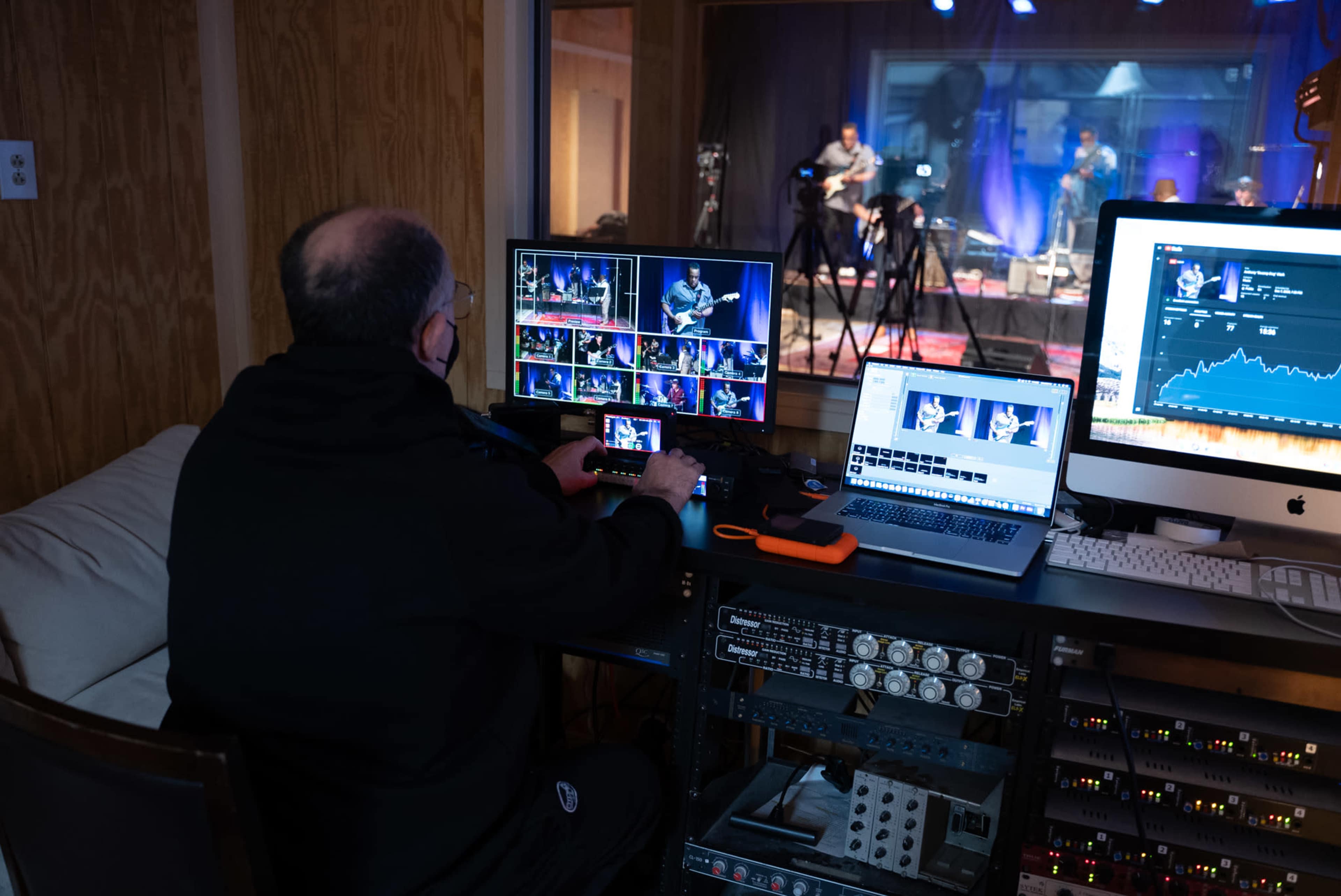 A technician operates video equipment in a control room while a band performs on stage in the background.