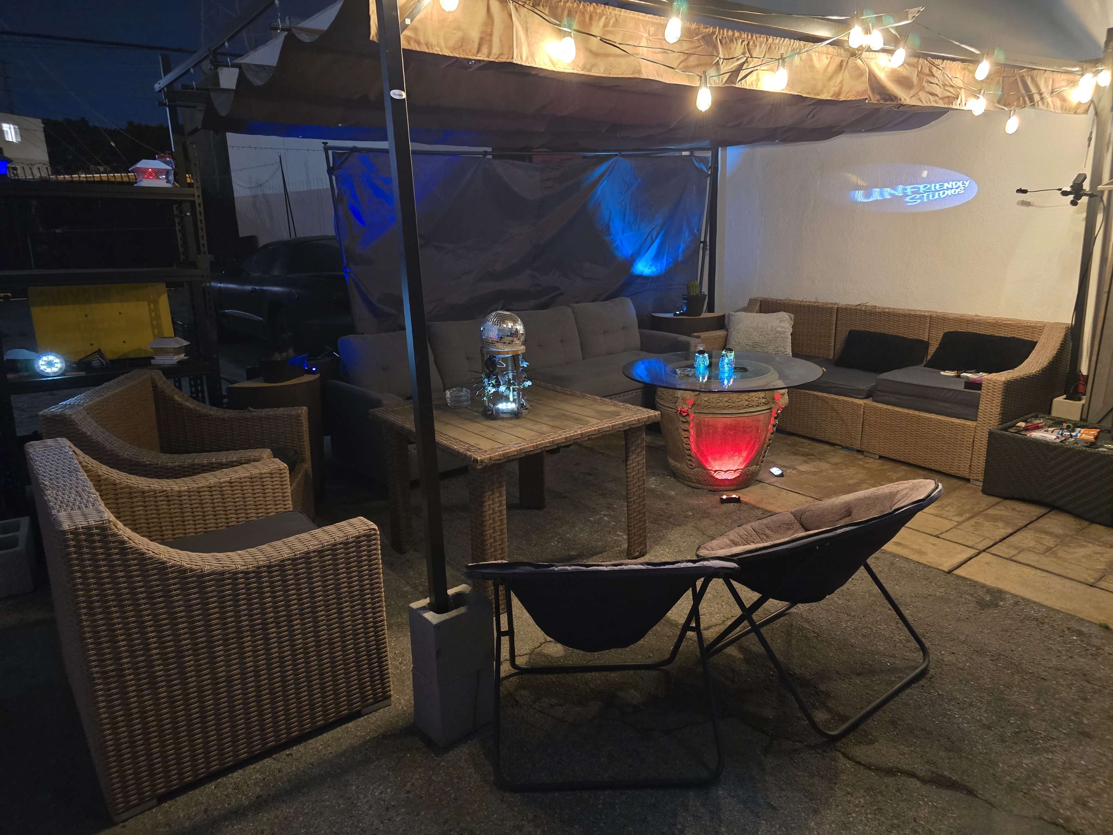 The image shows a cozy outdoor seating area featuring a variety of furniture, including a gray couch, wicker chairs, and a glass table with colorful lighting.