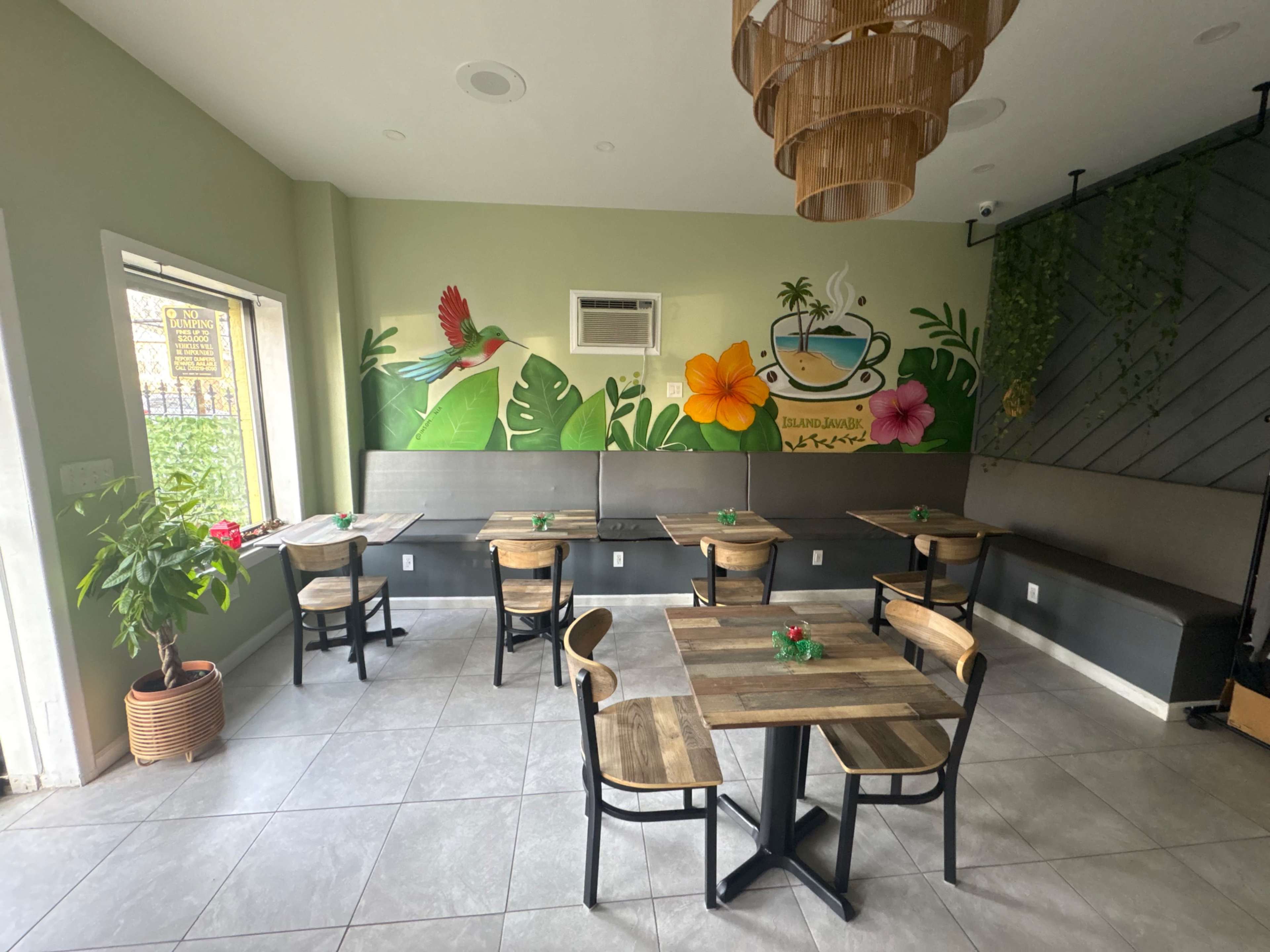 The image depicts a cozy café interior featuring wooden tables and chairs, with a vibrant tropical mural on the wall and plants decorating the space.