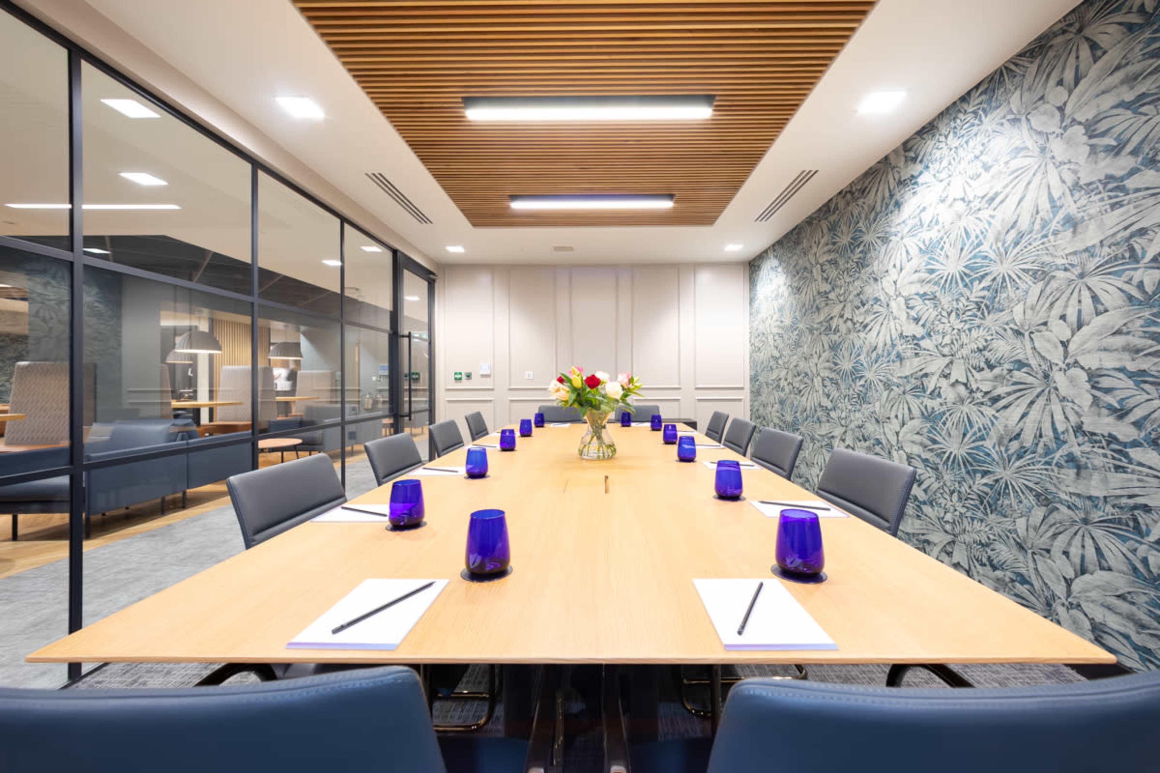 A modern conference room features a long wooden table with blue glass candle holders and notepads, surrounded by leather chairs and decorated with a floral centerpiece against a patterned wall.