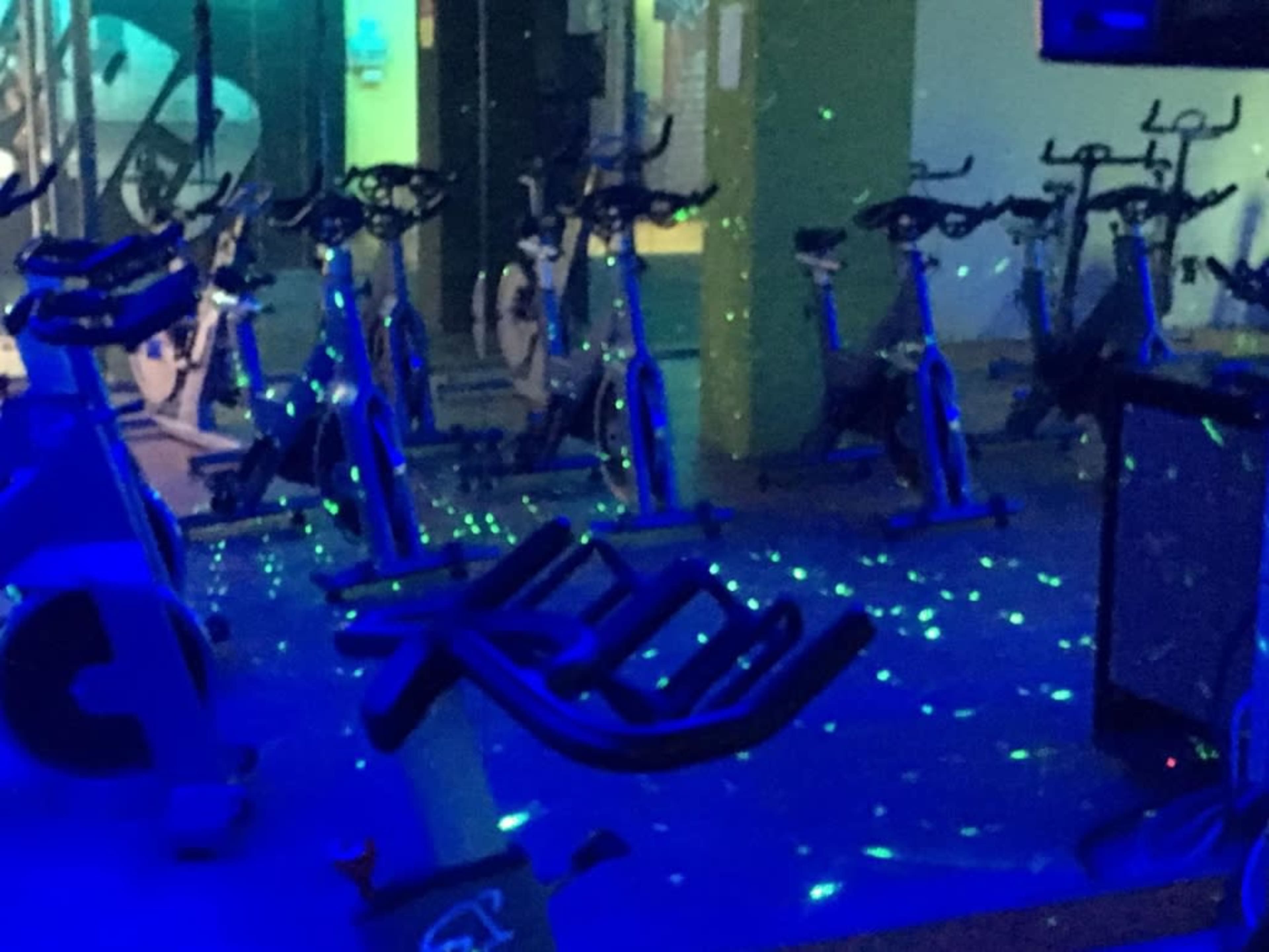 The image shows a fitness studio with stationary bikes arranged in a dimly lit space, illuminated by blue lights and green laser dots on the floor.