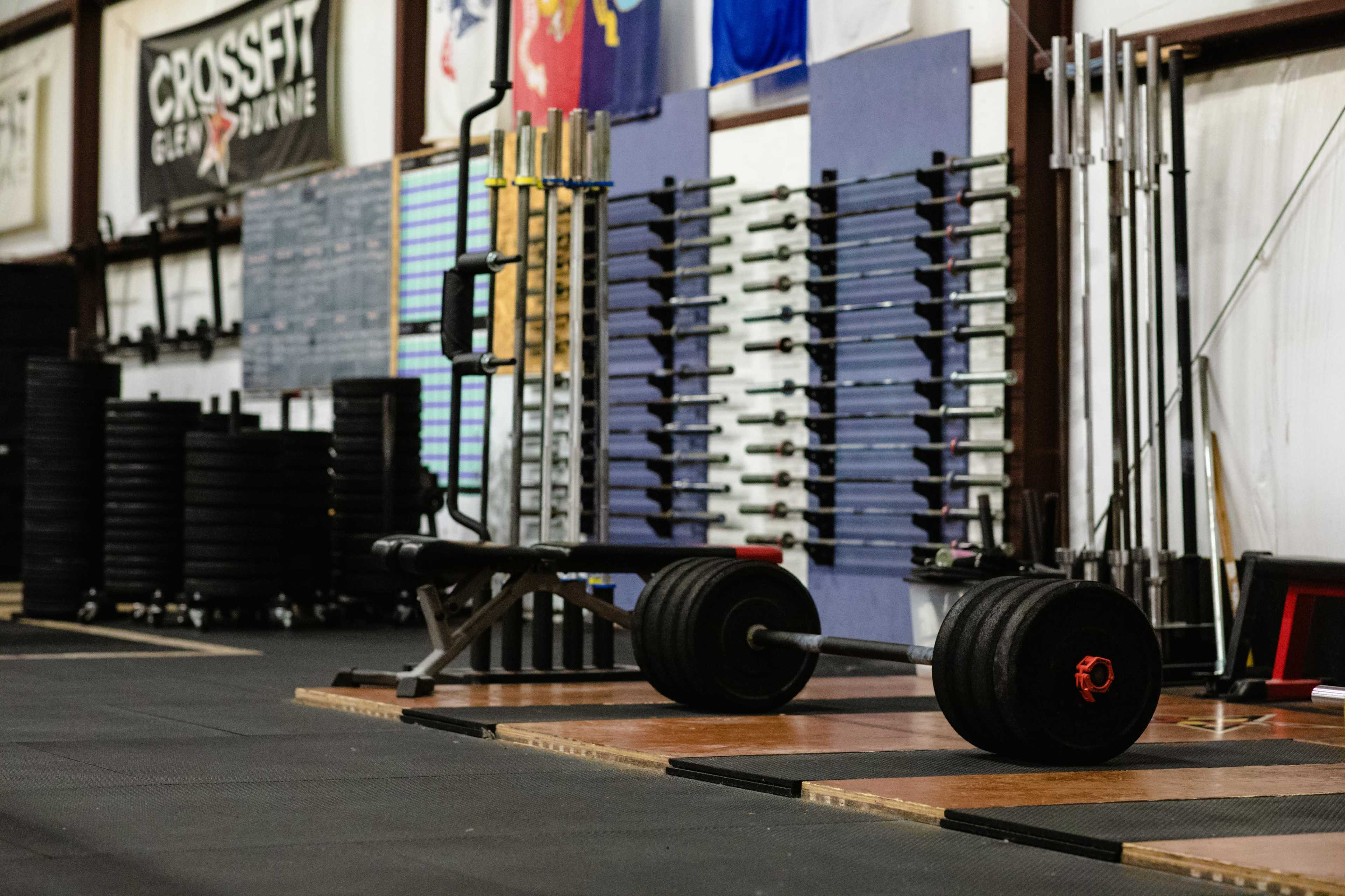 The image shows a CrossFit gym with weightlifting equipment, including a barbell and plates, arranged on a rubber floor.