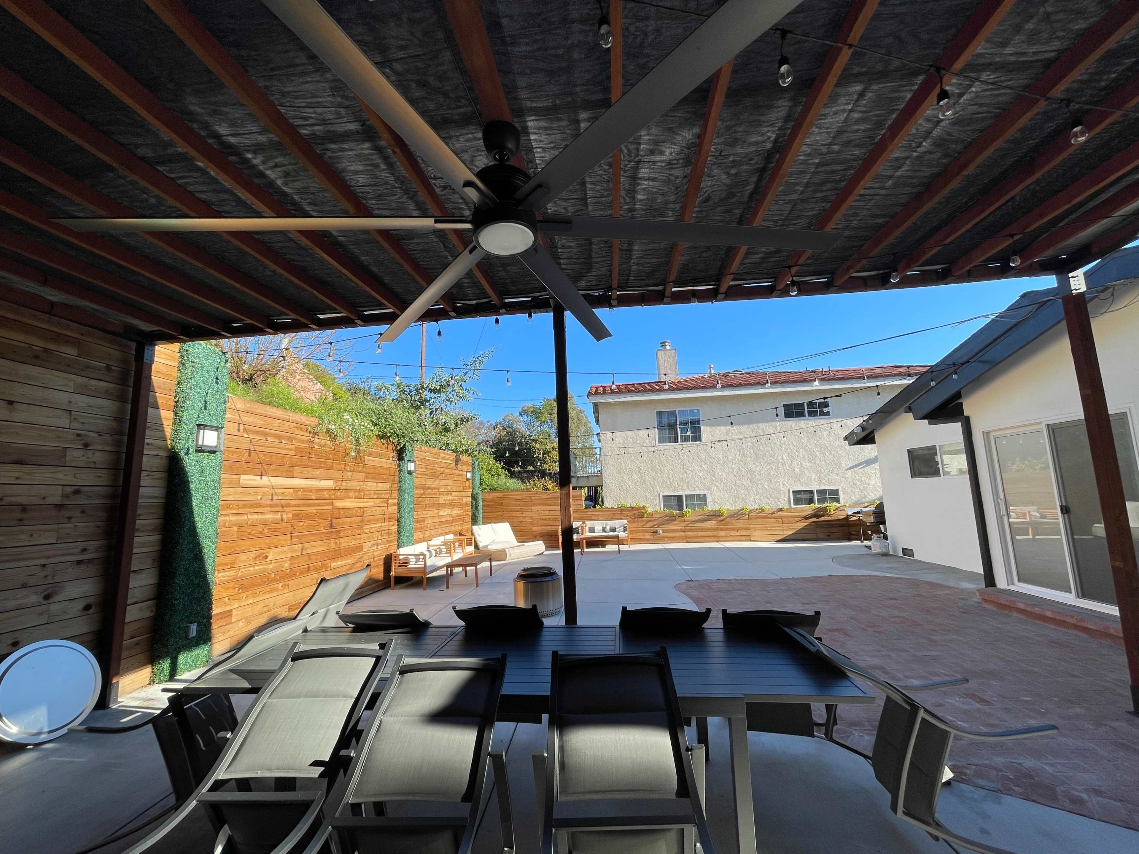 The image shows a covered outdoor patio area with a ceiling fan, a table surrounded by chairs, and a view of a landscaped backyard featuring a paved surface and wooden fencing.