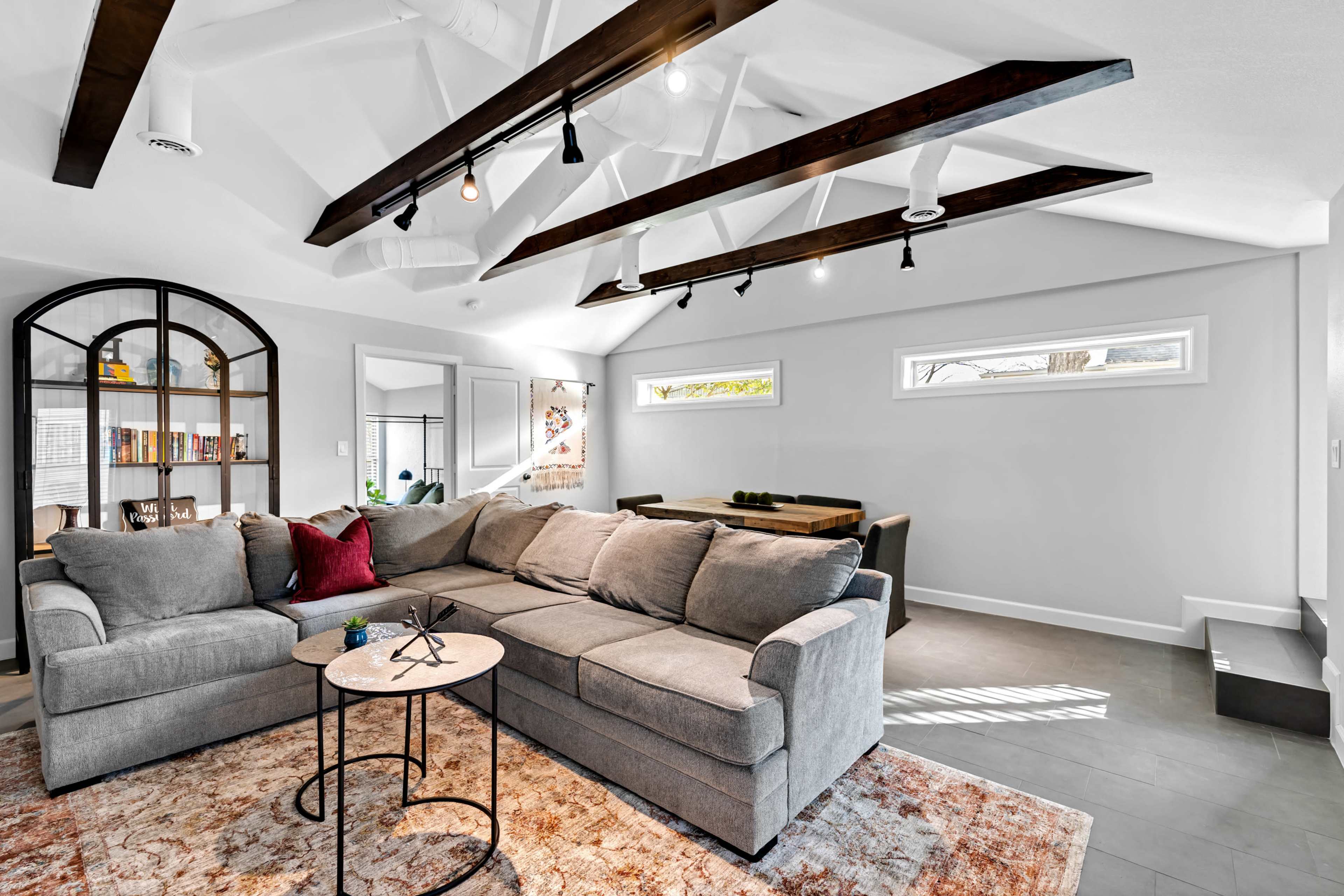 A modern living room featuring a gray sectional sofa, a round side table, a bookshelf, and a dining area, all under a ceiling with exposed beams and large windows.