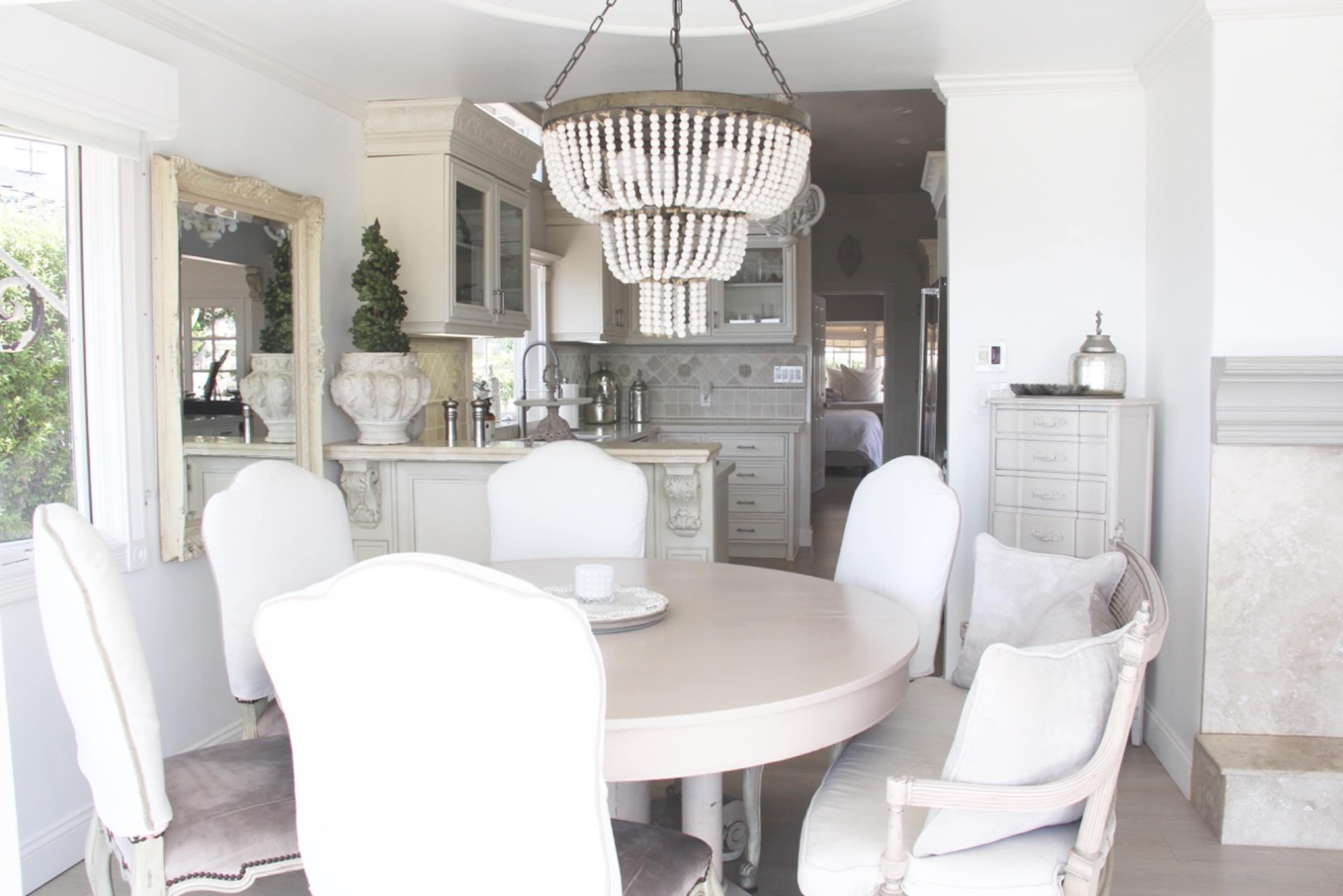 A dining area features a round table surrounded by upholstered chairs and a beaded chandelier, with a kitchen visible in the background.