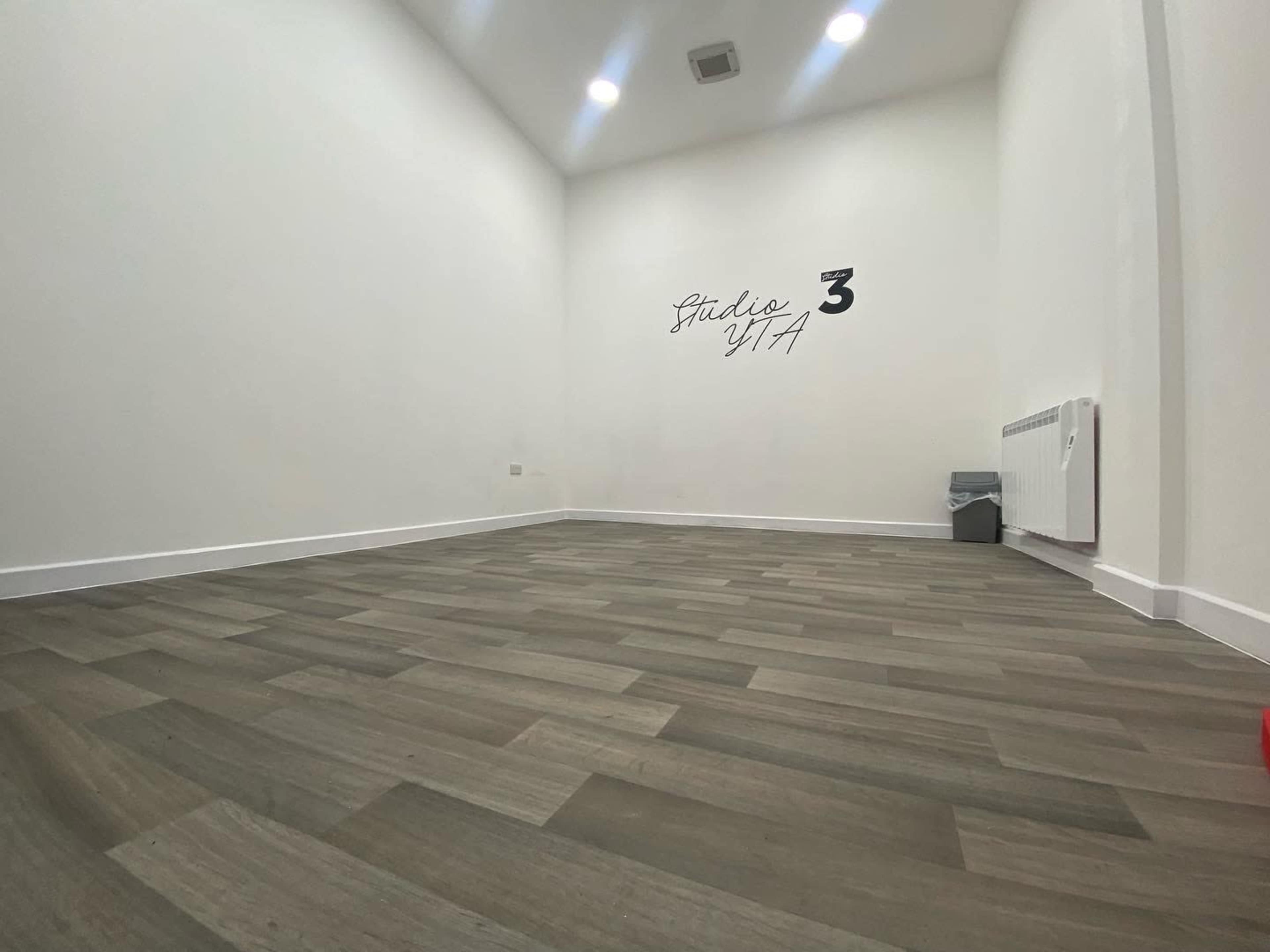 A spacious room with wooden flooring, white walls, and minimal furnishings, including a heater and a trash can.