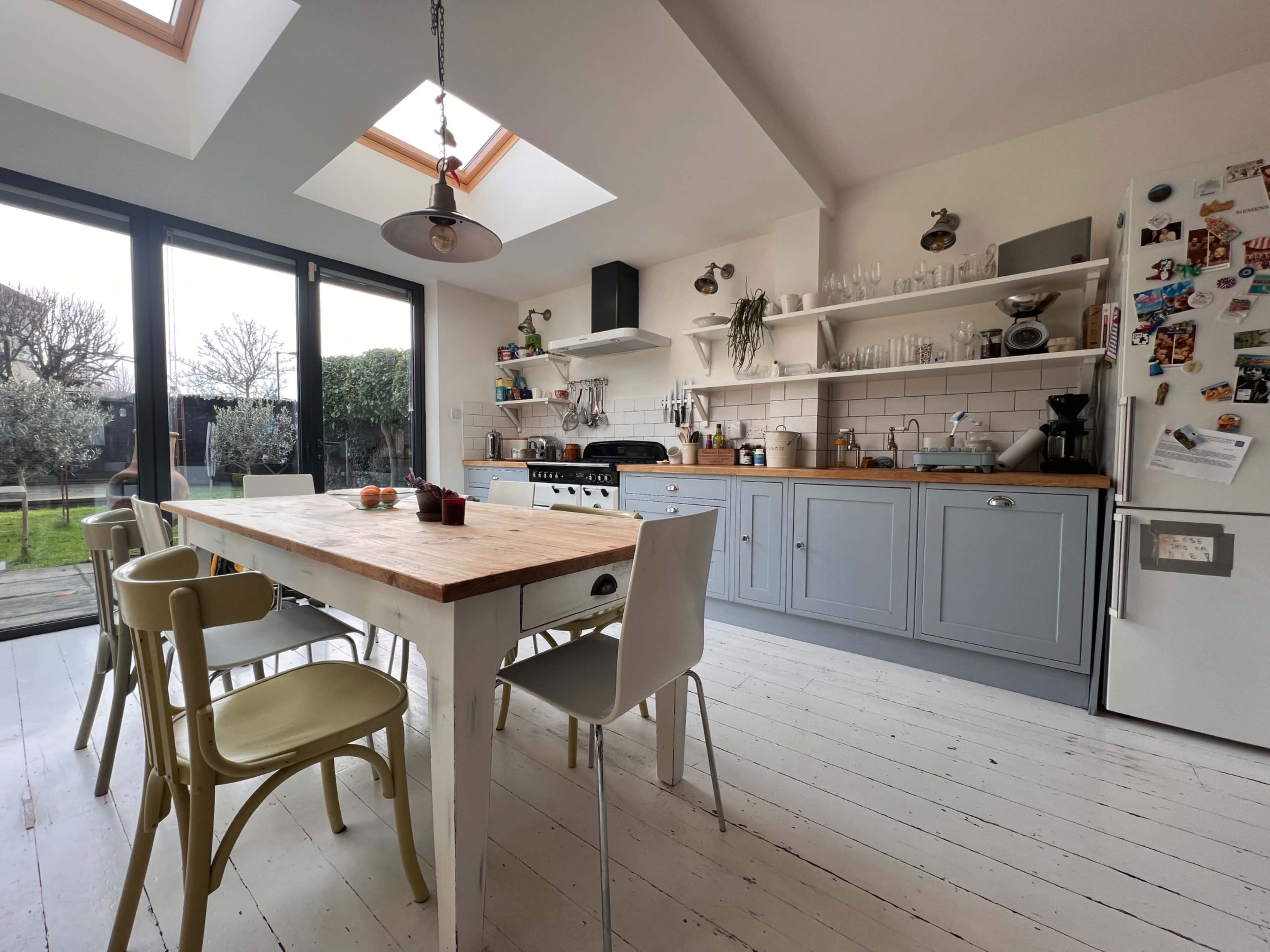 A modern kitchen features a large wooden dining table surrounded by chairs, with light streaming in through skylights and large glass doors opening to a garden.