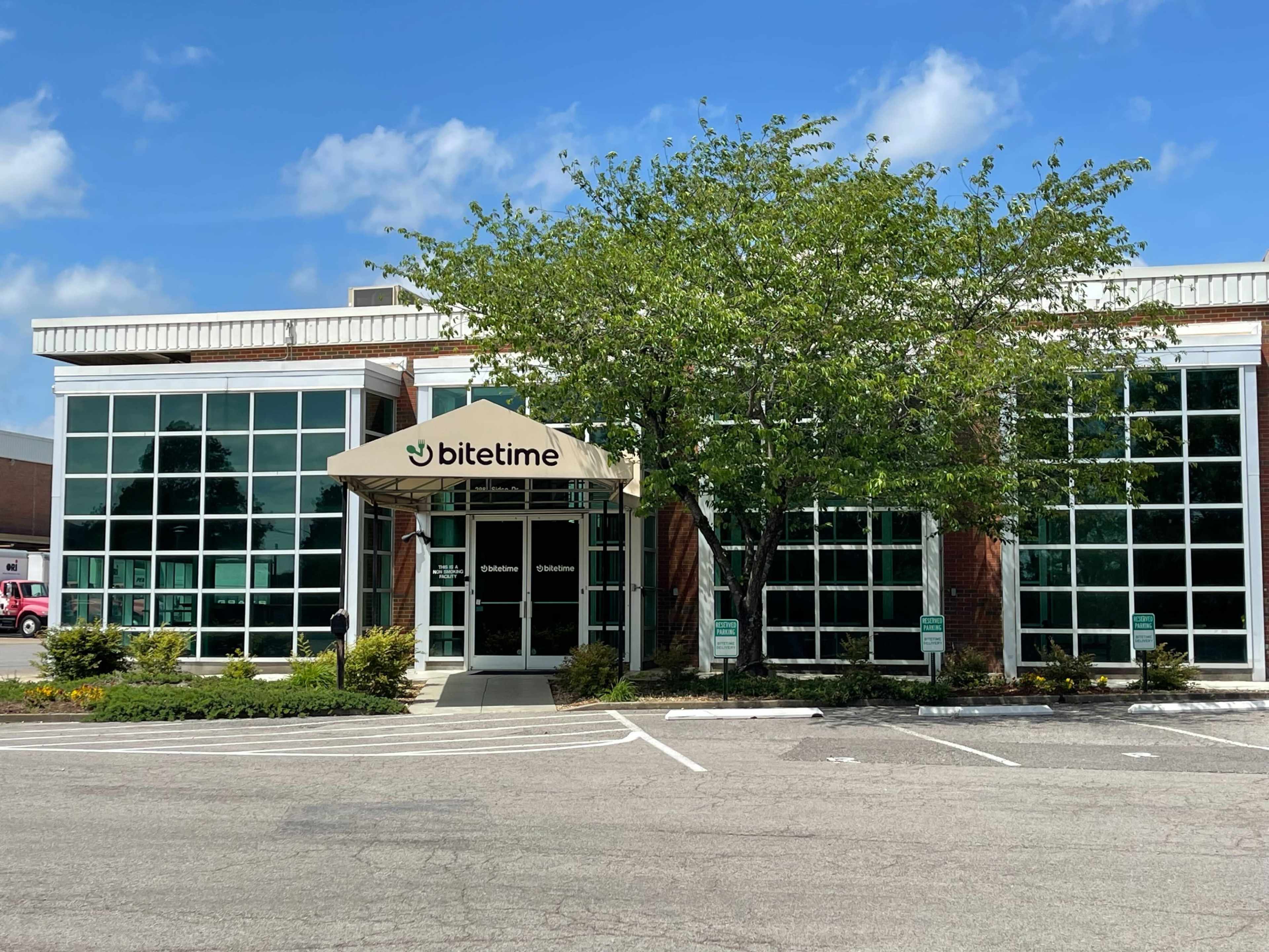The image shows a modern building with large windows and a canopy labeled "bitetime" at the entrance, surrounded by well-maintained landscaping.