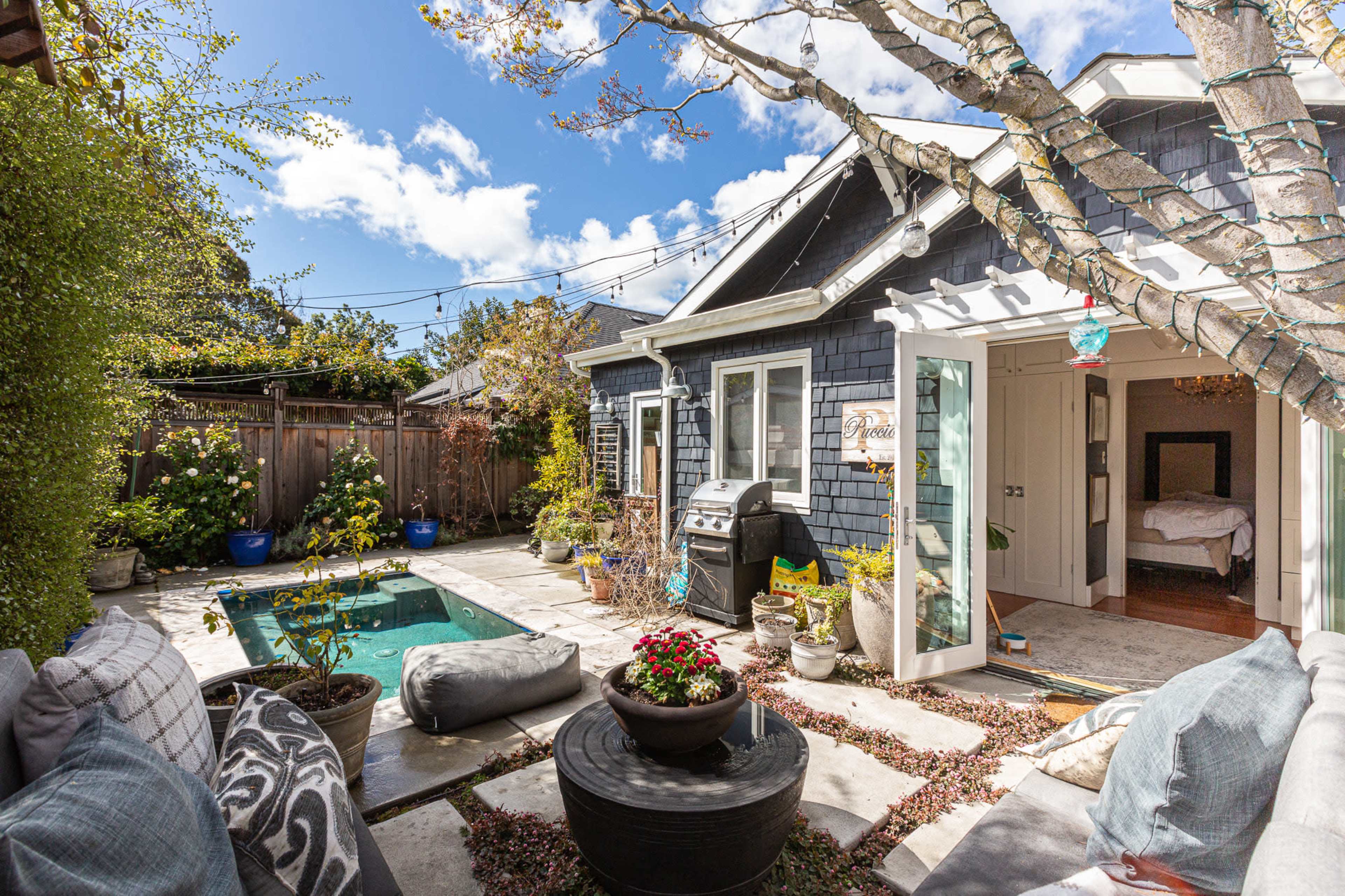 A cozy backyard features a small pool, patio furniture, and a grill, with a house in the background and trees overhead.