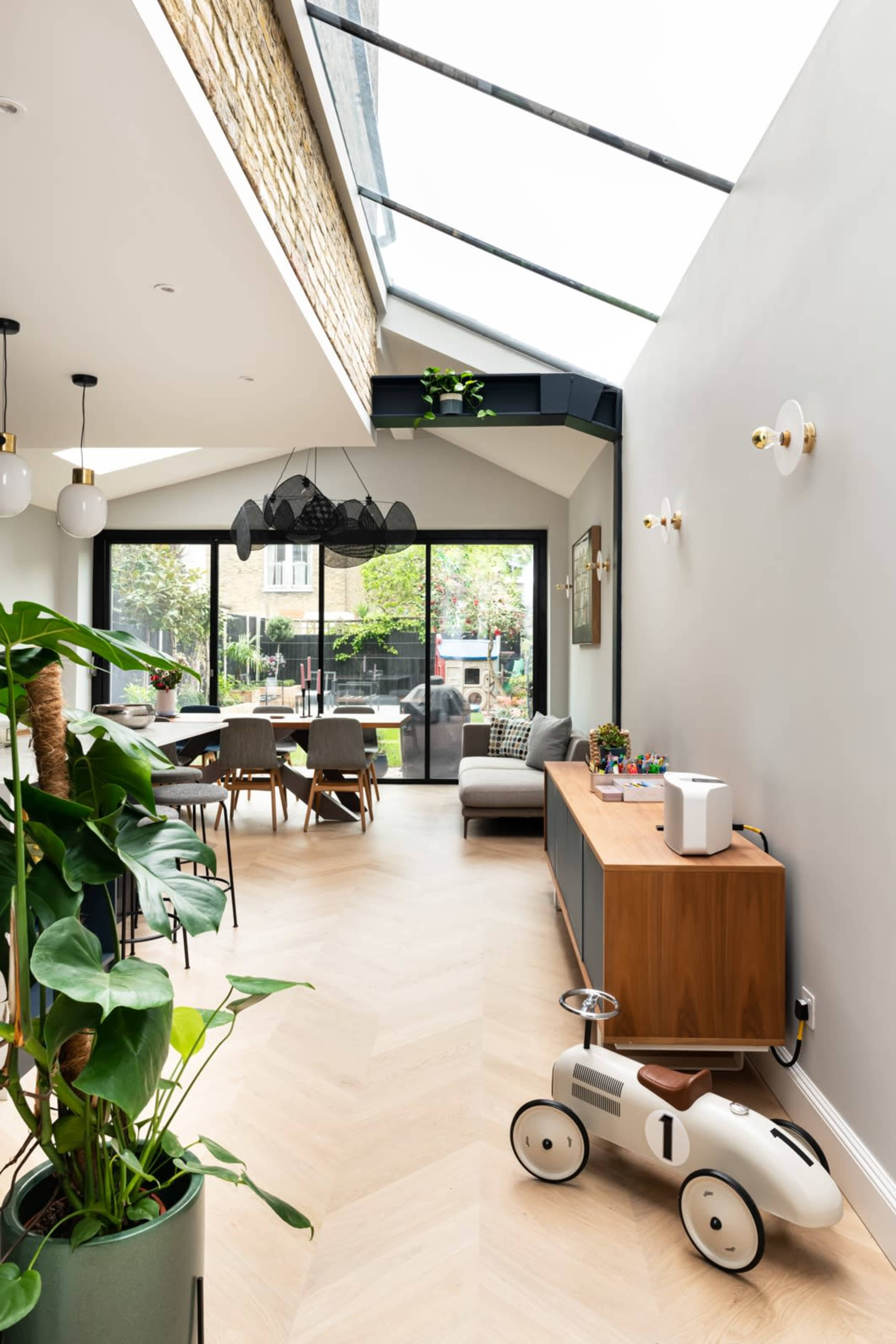 The image shows a modern living area with a wooden sideboard, a children's toy car, and large glass doors leading to an outdoor space, illuminated by natural light from a skylight.
