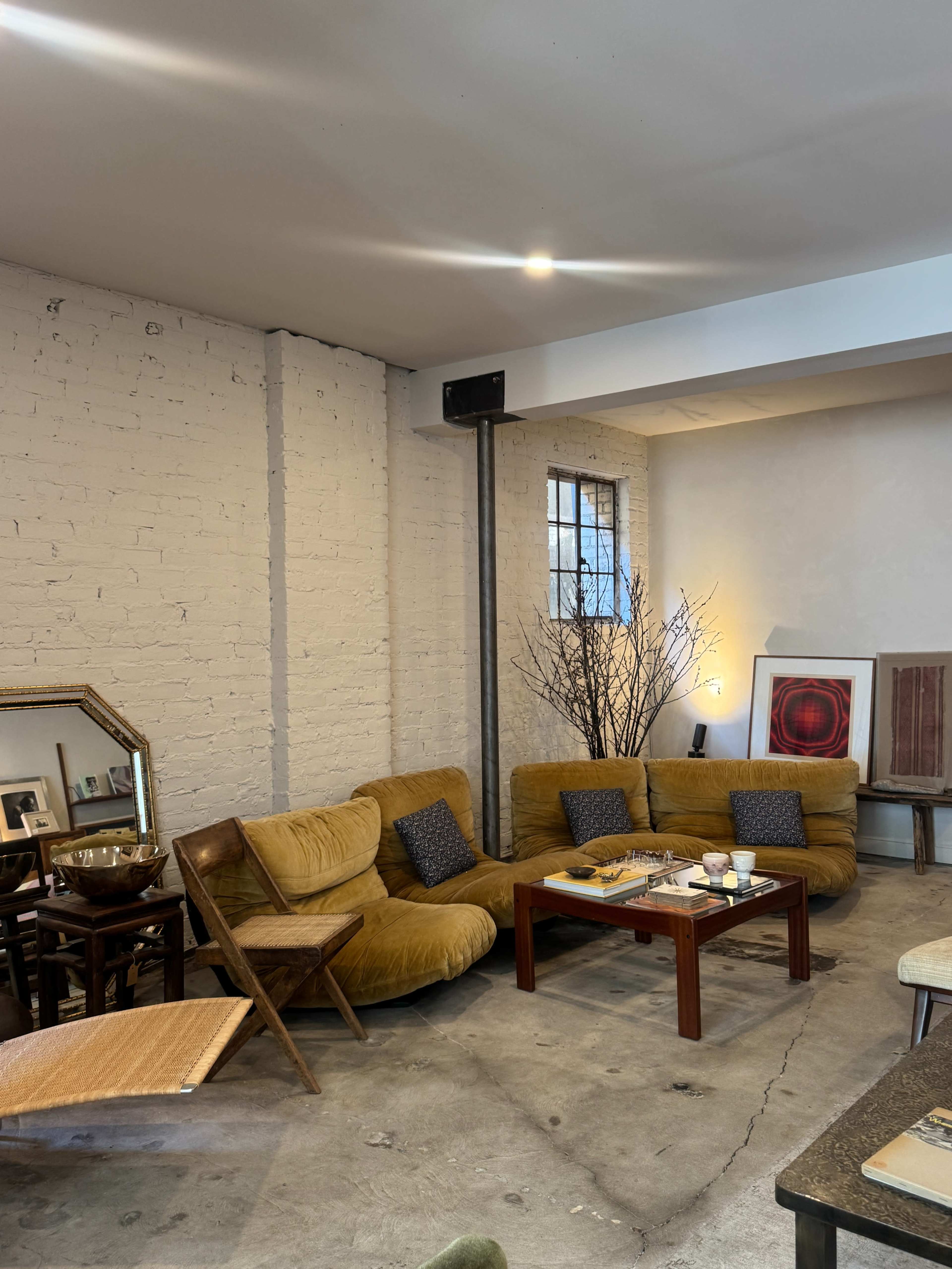 The image shows a cozy, modern living space featuring a large yellow sectional couch, a wooden coffee table, and a mix of decor elements, including a mirror and decorative branches.
