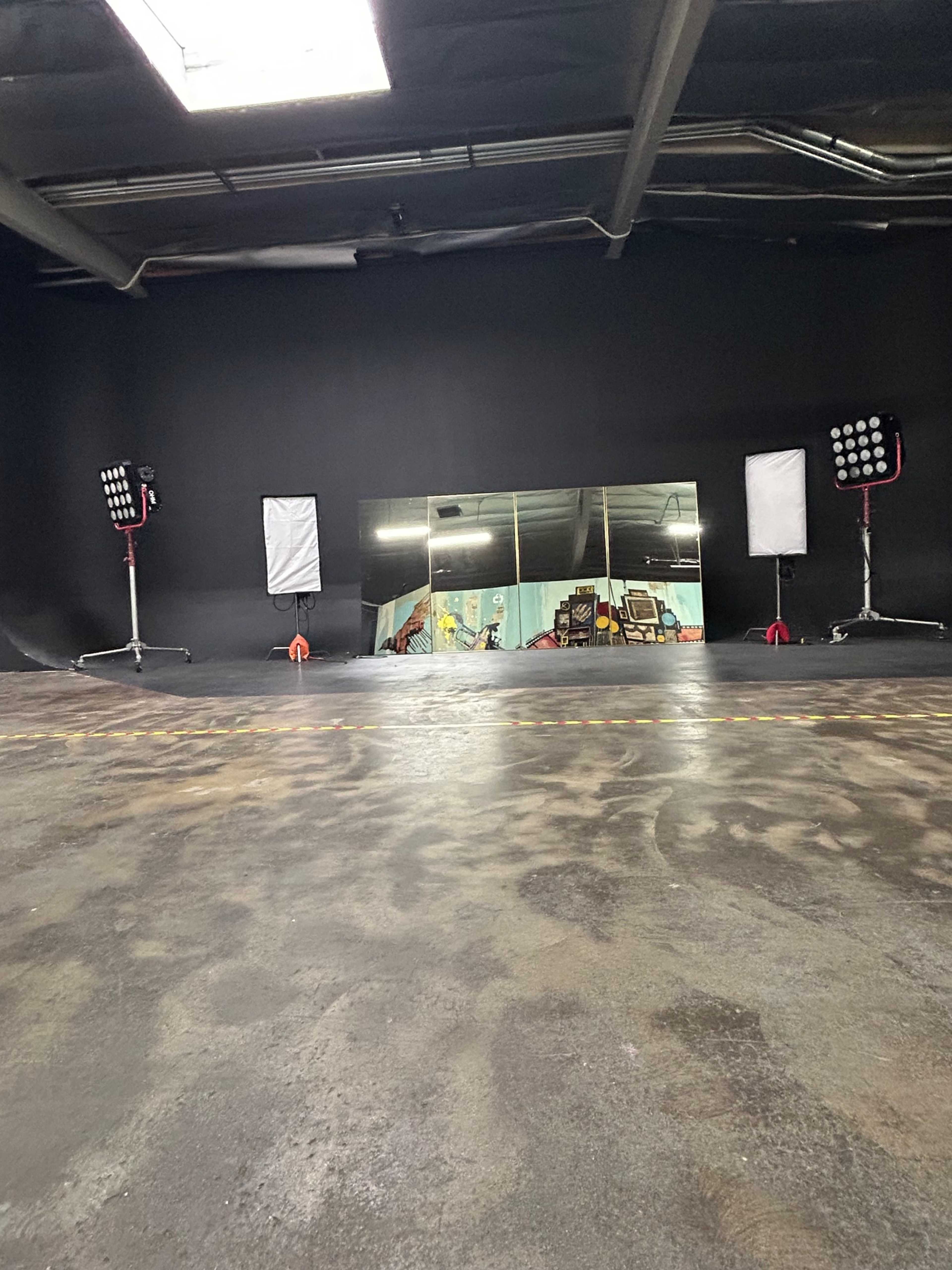 A large, black-walled studio space features a wide mirror and two sets of bright lights on stands, with a concrete floor visible in the foreground.