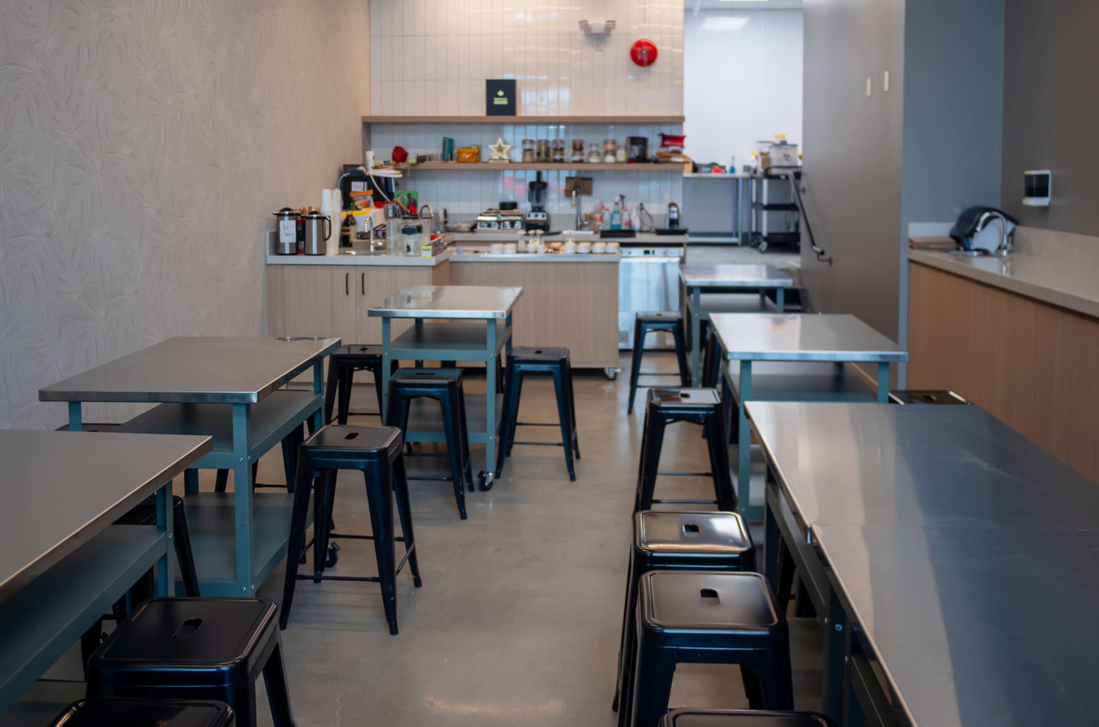 The image shows a modern kitchen workspace with metal tables and stools arranged in rows, featuring various kitchen appliances and supplies on a countertop.