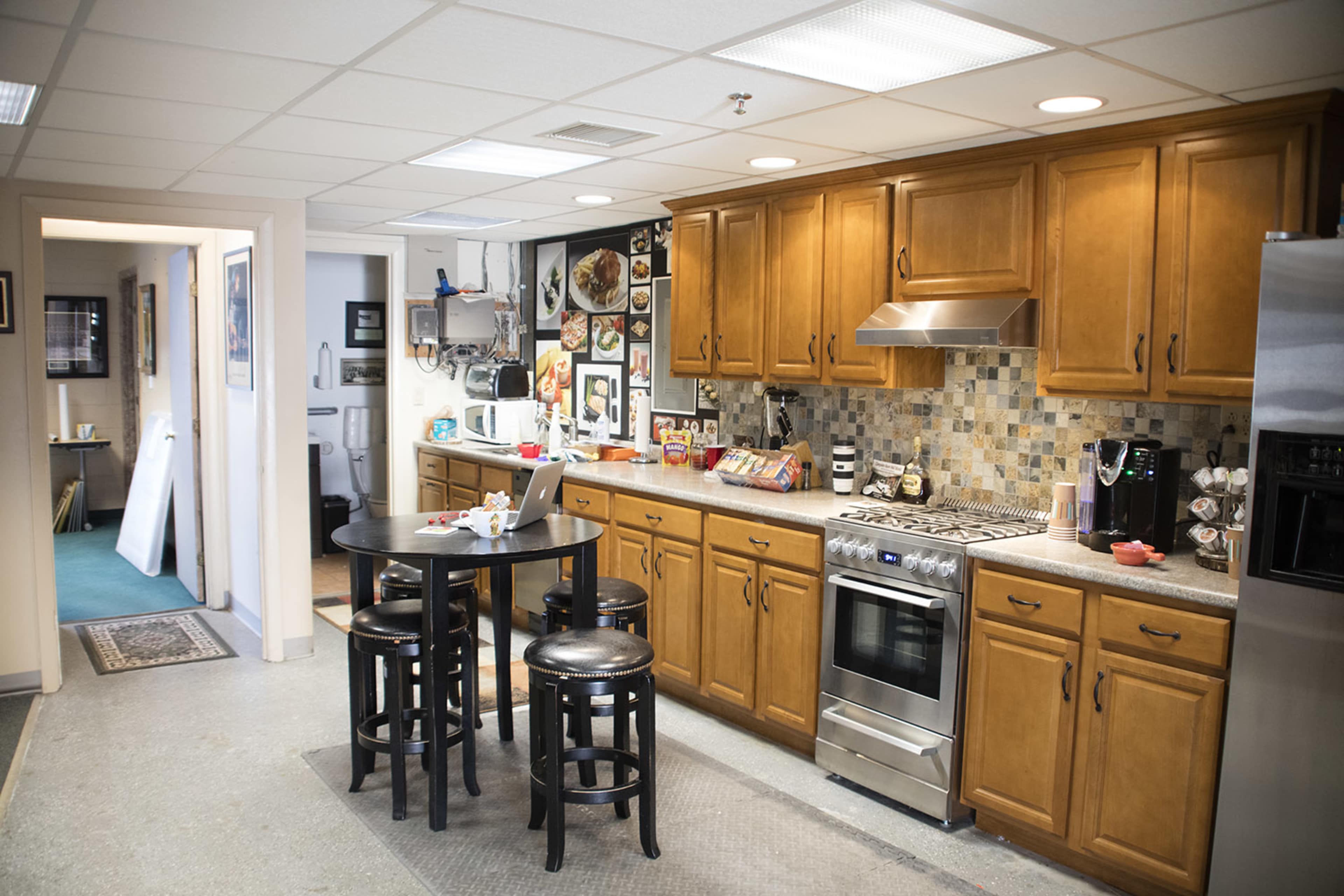 A modern kitchen with wooden cabinets, a countertop, a stove, and a table with black stools, adjacent to a hallway leading to another room.