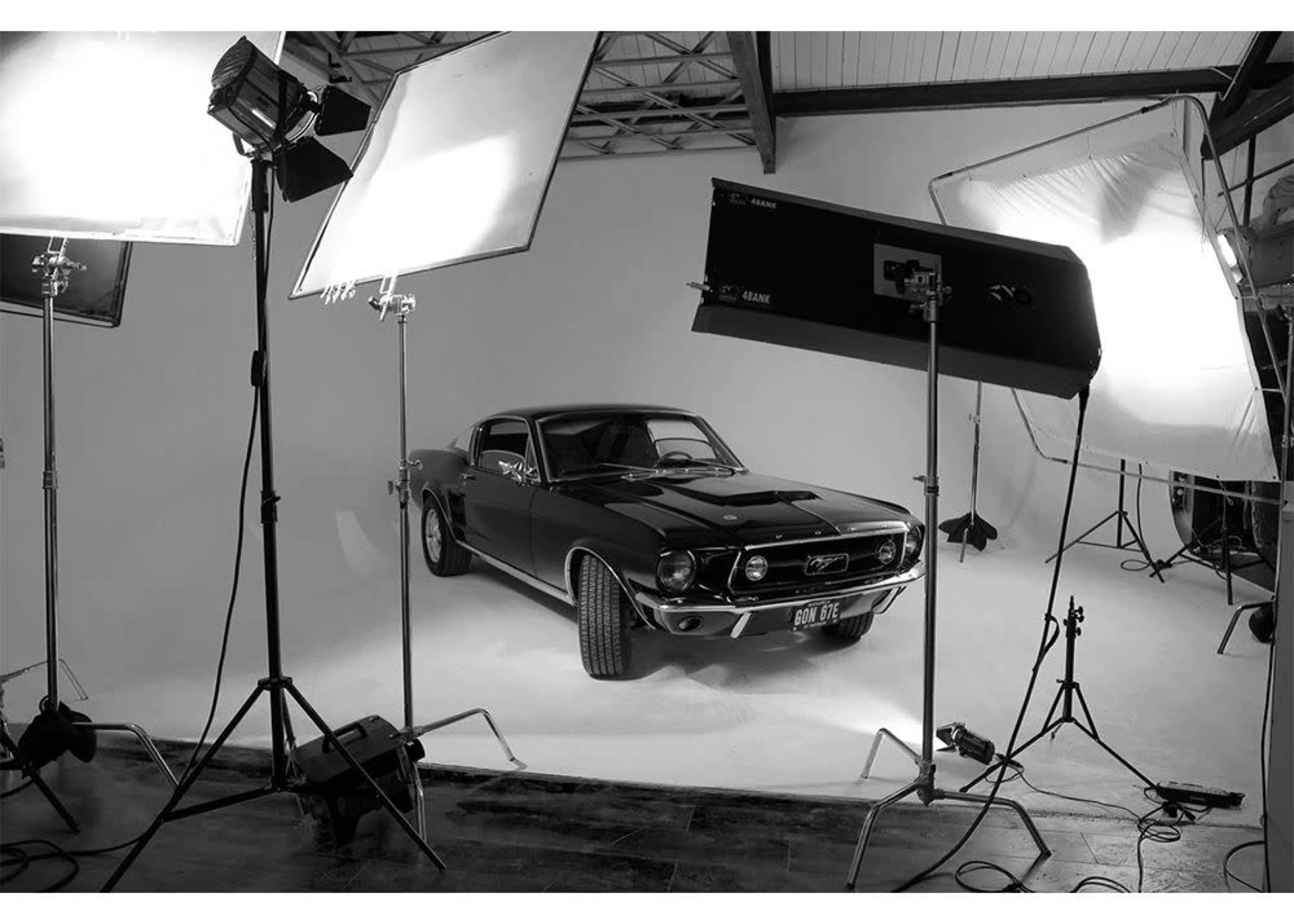 A classic black Mustang is positioned in a studio setting surrounded by multiple lighting equipment.