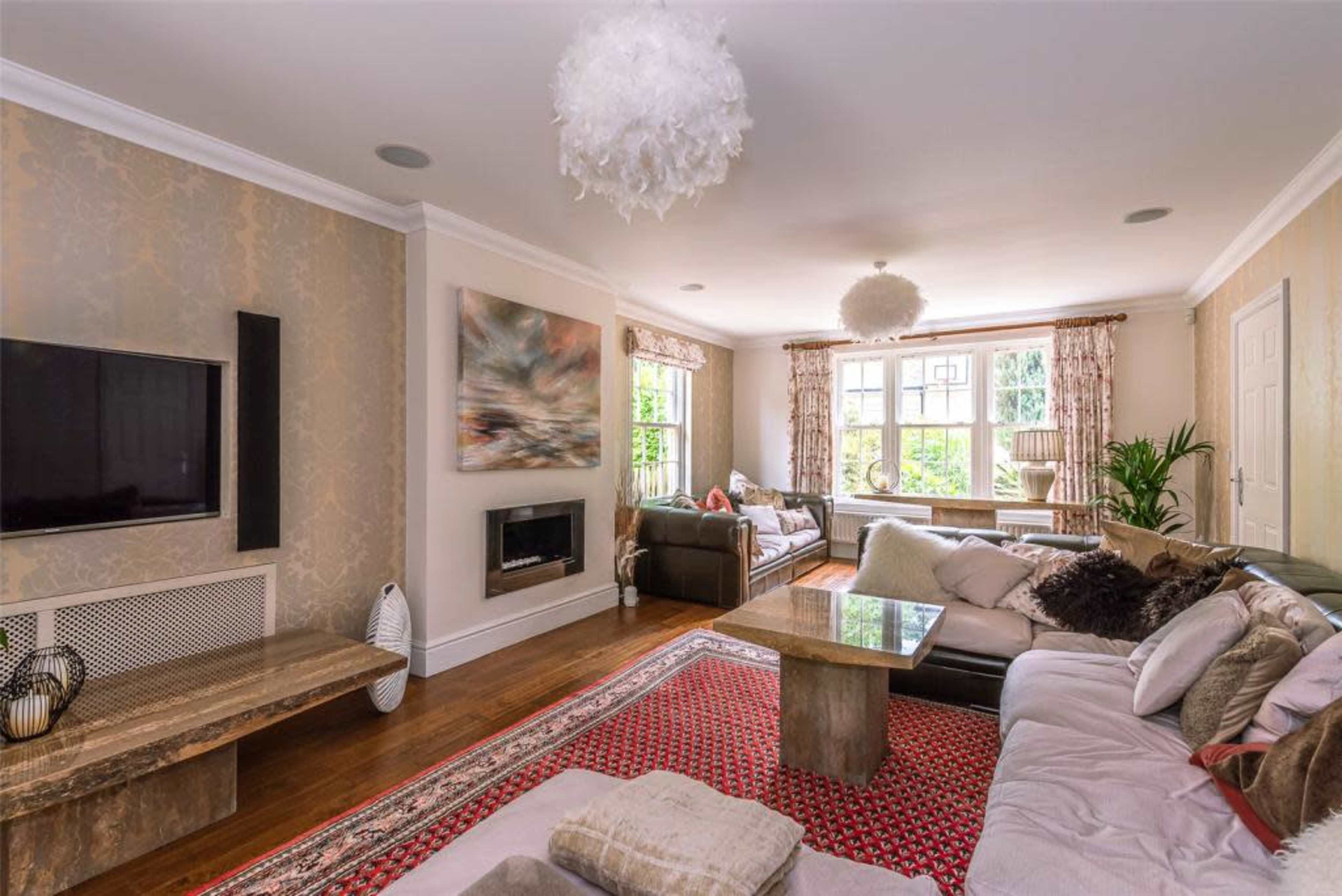 The living room features a large area rug, multiple couches, a mounted television, and a fireplace, with natural light streaming in through large windows.