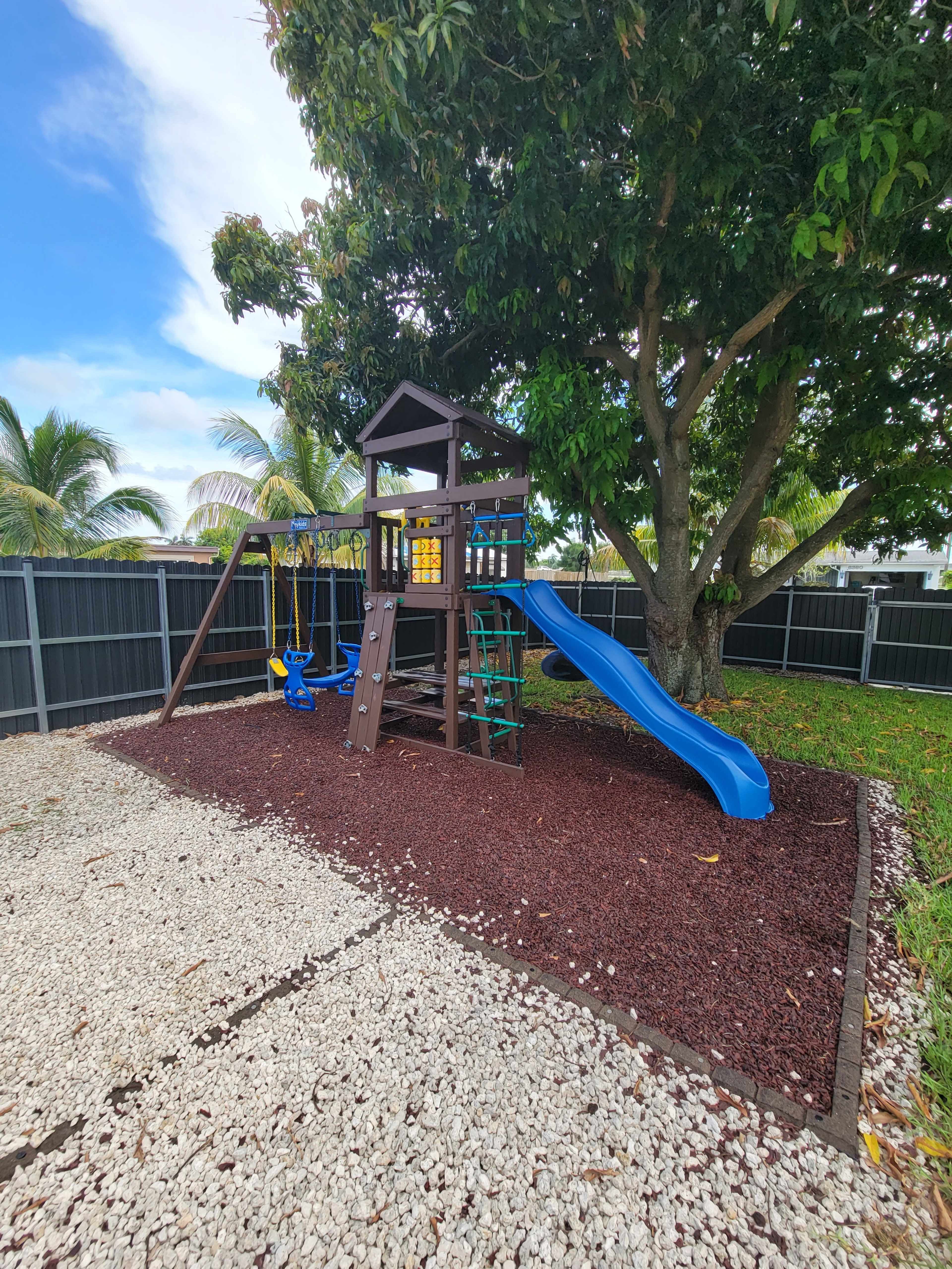 The image shows a wooden playground set with a slide, swings, and climbing features, situated in a landscaped yard with gravel and surrounded by a black fence and palm trees.