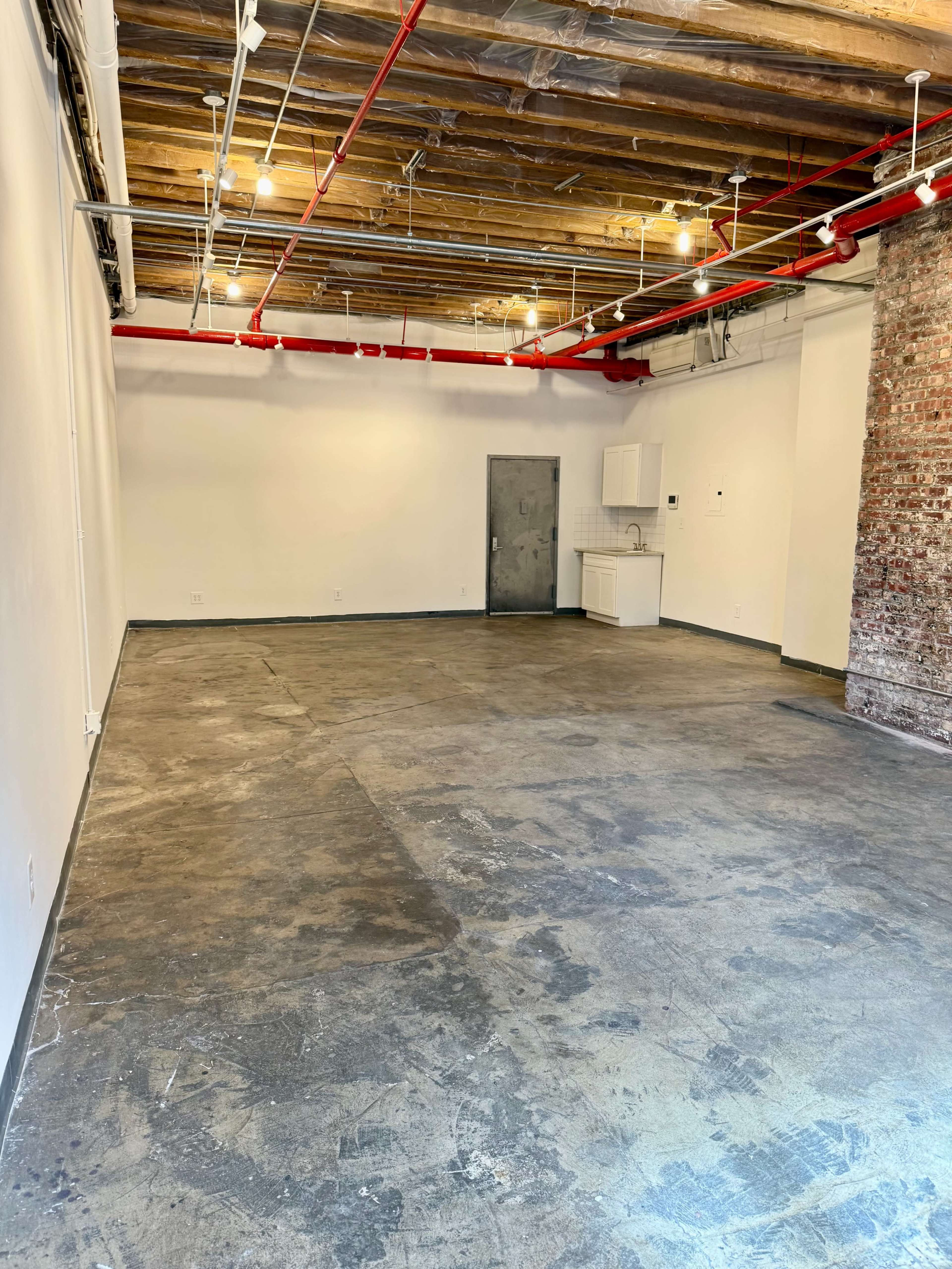 The image shows an empty room with a concrete floor, exposed wooden beams, a red pipe system, and a kitchenette along one wall.