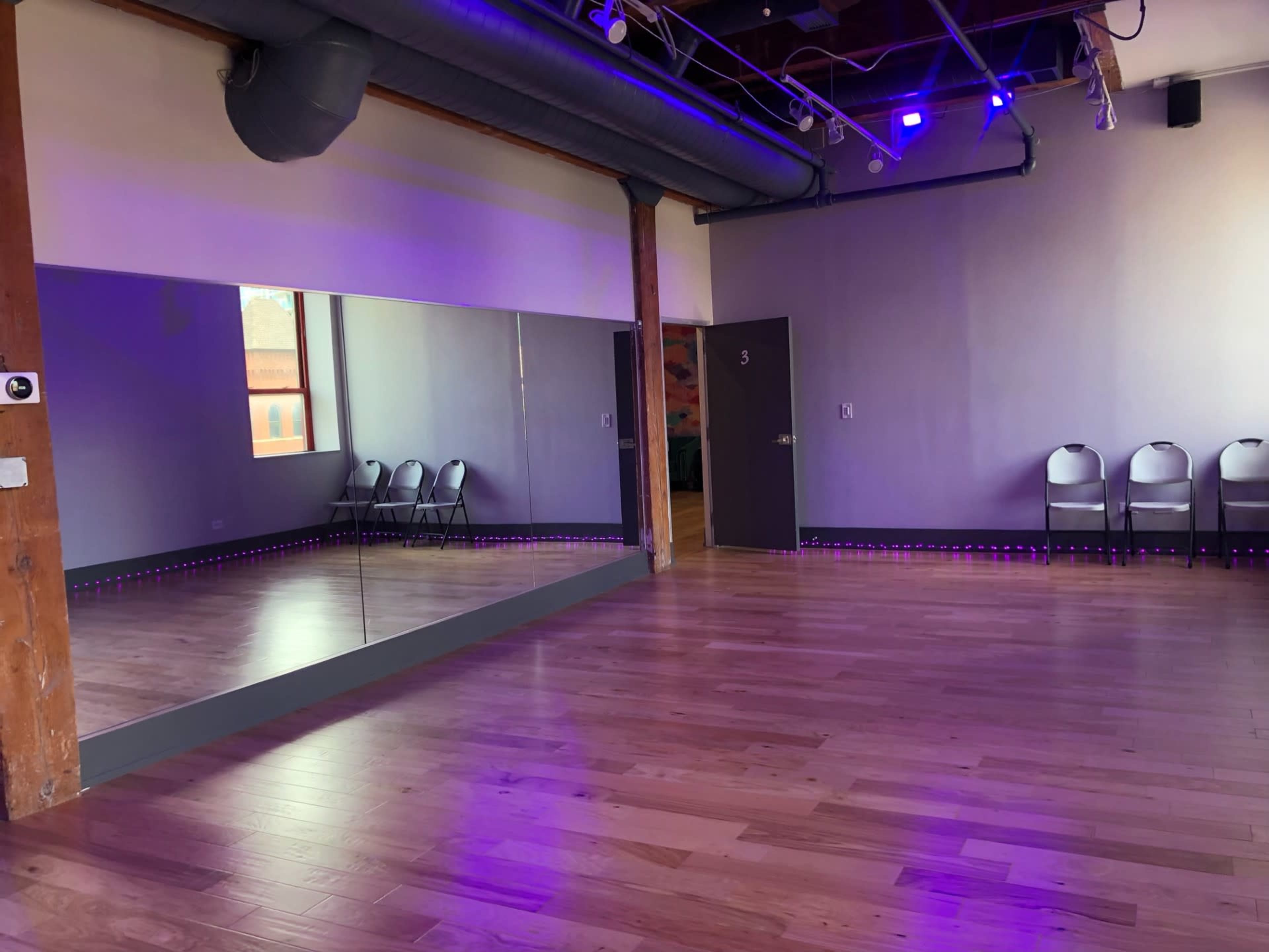 A dance studio with hardwood flooring, a large mirror on one wall, and several folding chairs arranged against a side wall under purple lighting.